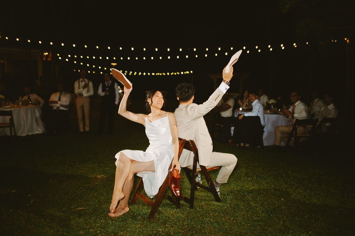 Bride and groom playing shoe game on chairs under string lights at an outdoor evening wedding reception.
