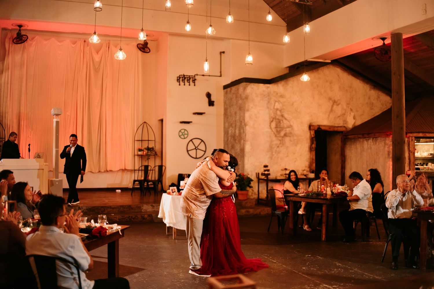 Couple shares first dance in rustic wedding venue with warm Edison bulb lighting and guests seated at tables.