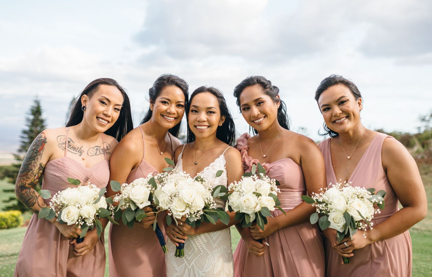 Bridal party in pink dresses holds white flower bouquets while posing outdoors.