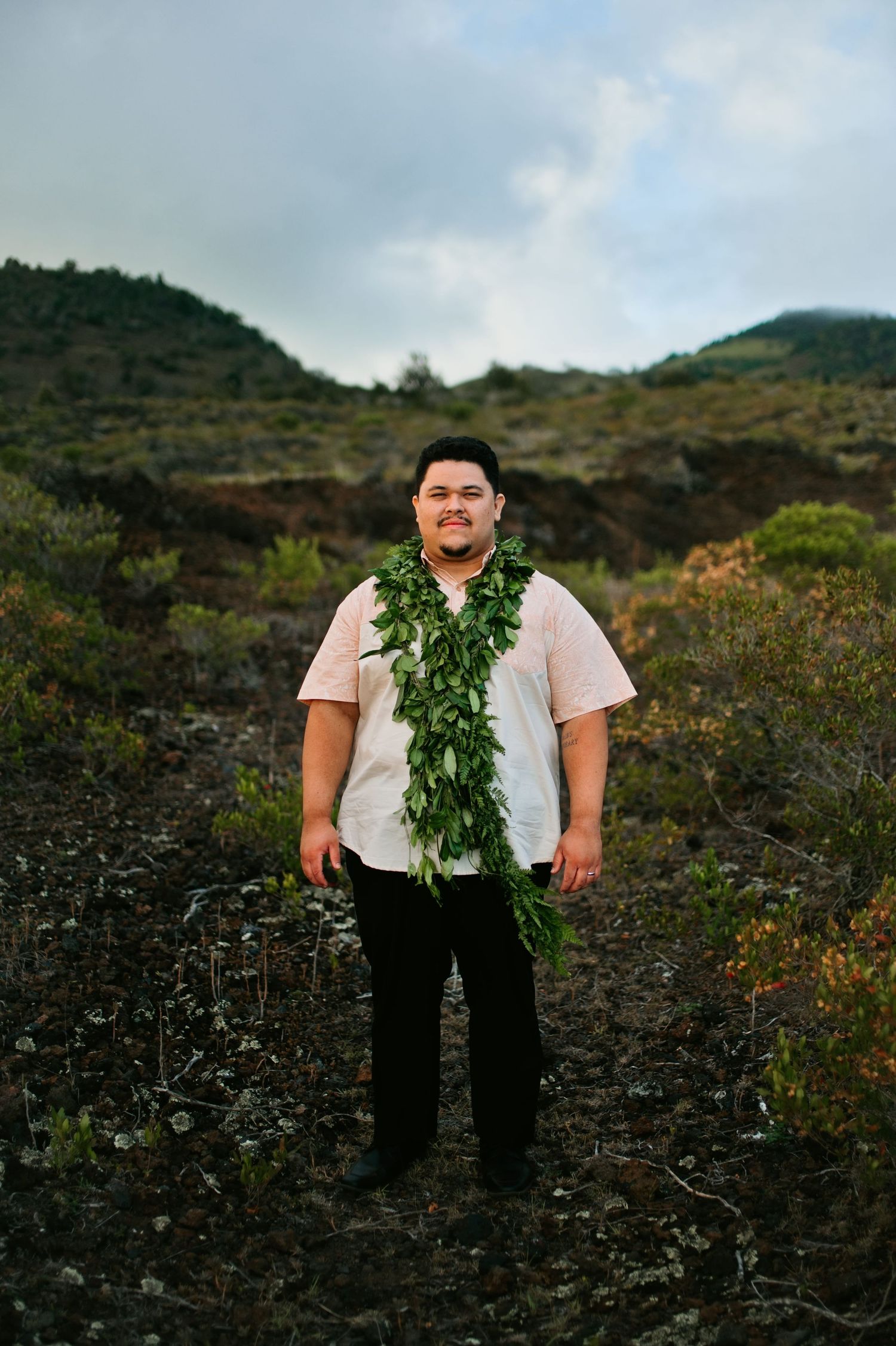 Man wearing a green leaf lei stands in a volcanic landscape with lush hills under a cloudy sky.