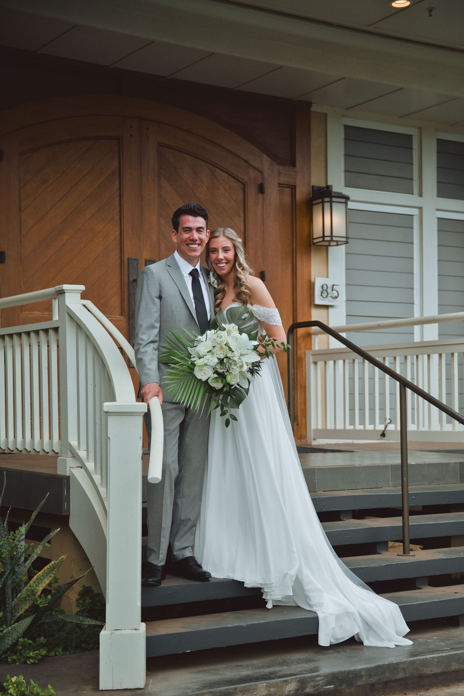 Bride and groom posing on church steps, bride holding tropical bouquet, groom in gray suit, building number 815.