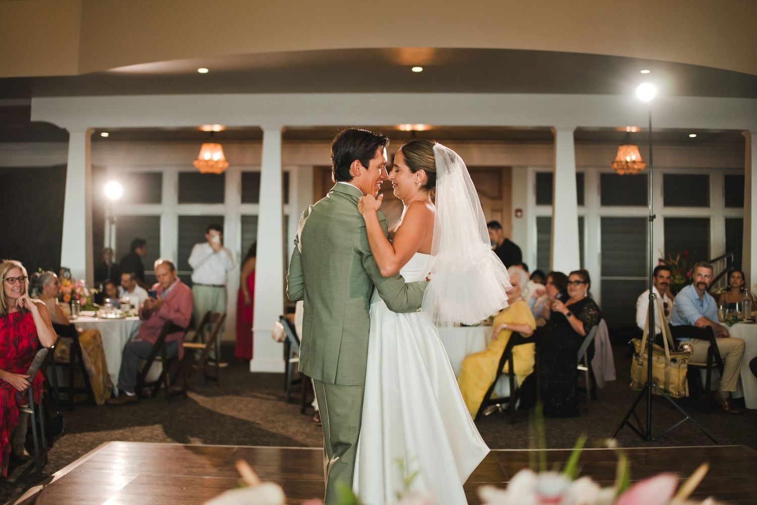 Bride and groom share first dance at elegant indoor wedding reception with guests seated at candlelit tables.