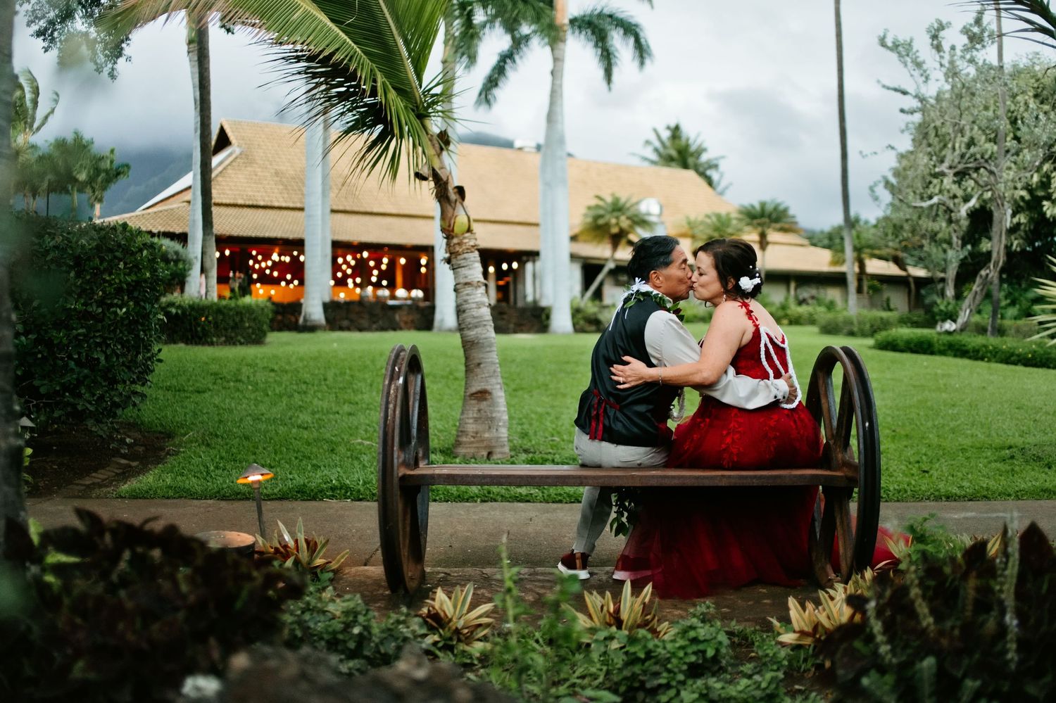 Couple sharing a romantic kiss on a vintage cart surrounded by lush tropical gardens at a Hawaiian resort.