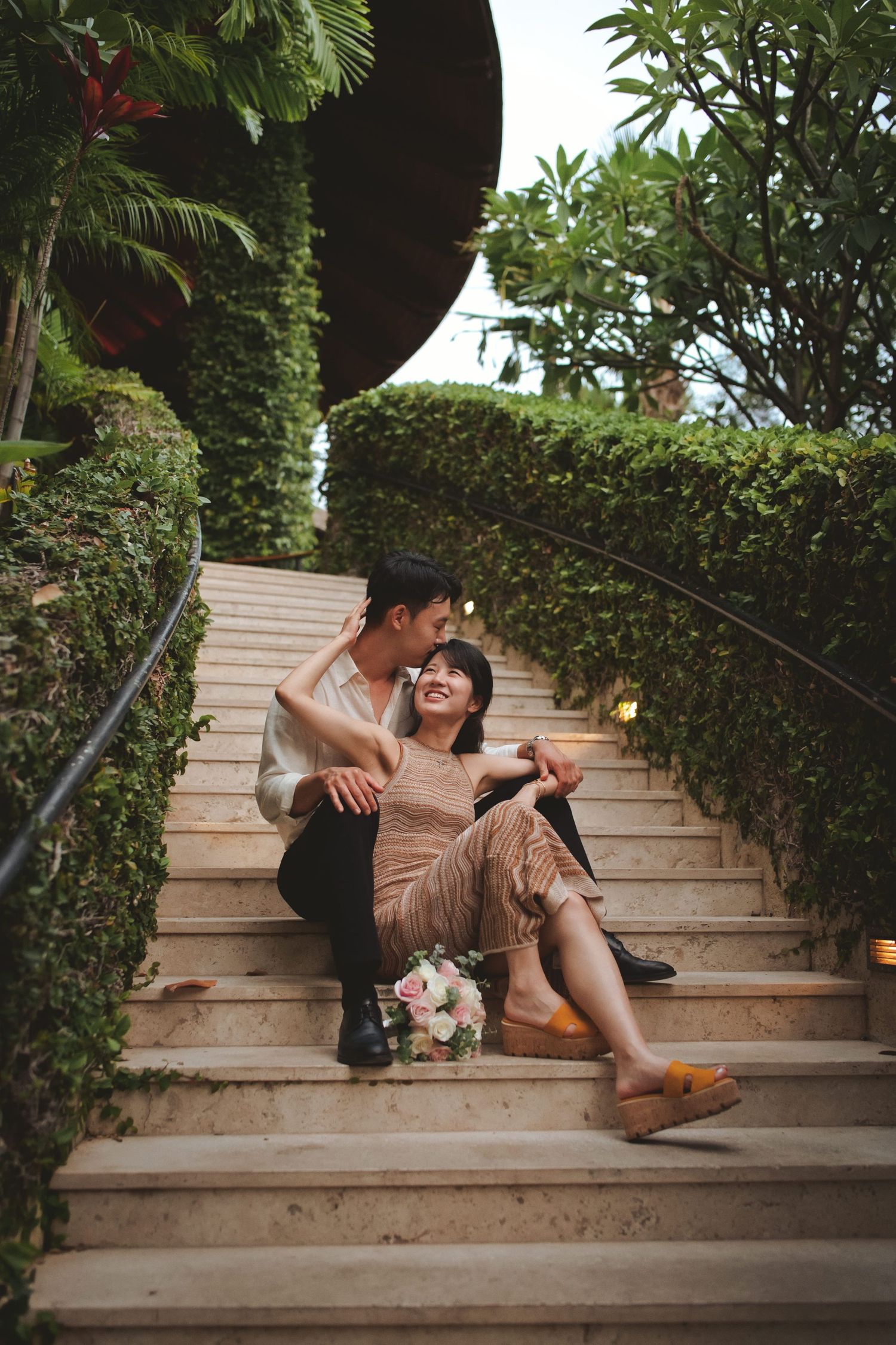 Couple sitting on outdoor stone steps surrounded by lush greenery, smiling at each other with a bouquet of flowers nearby.