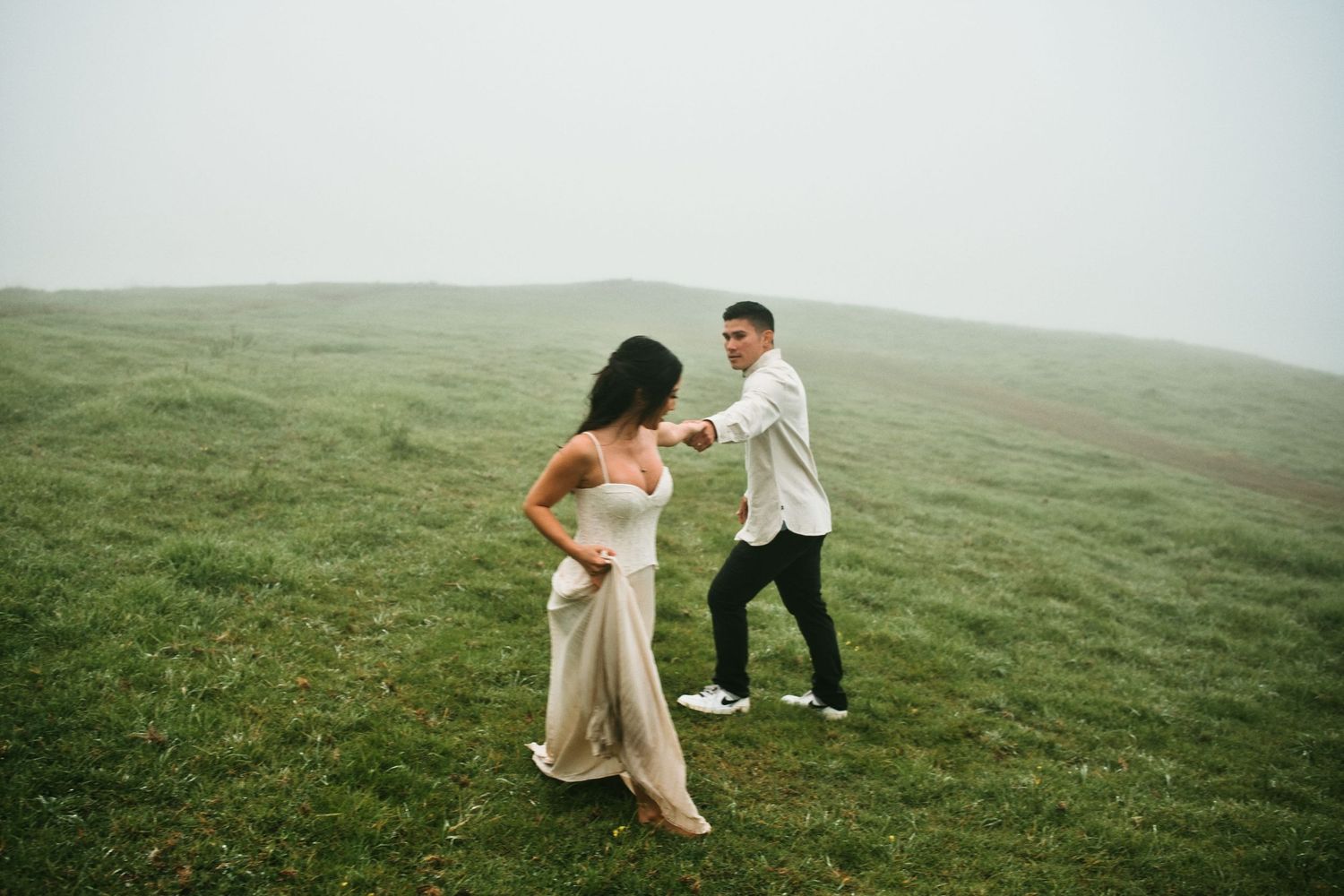 Couple dancing and holding hands on a foggy green hillside during engagement shoot.