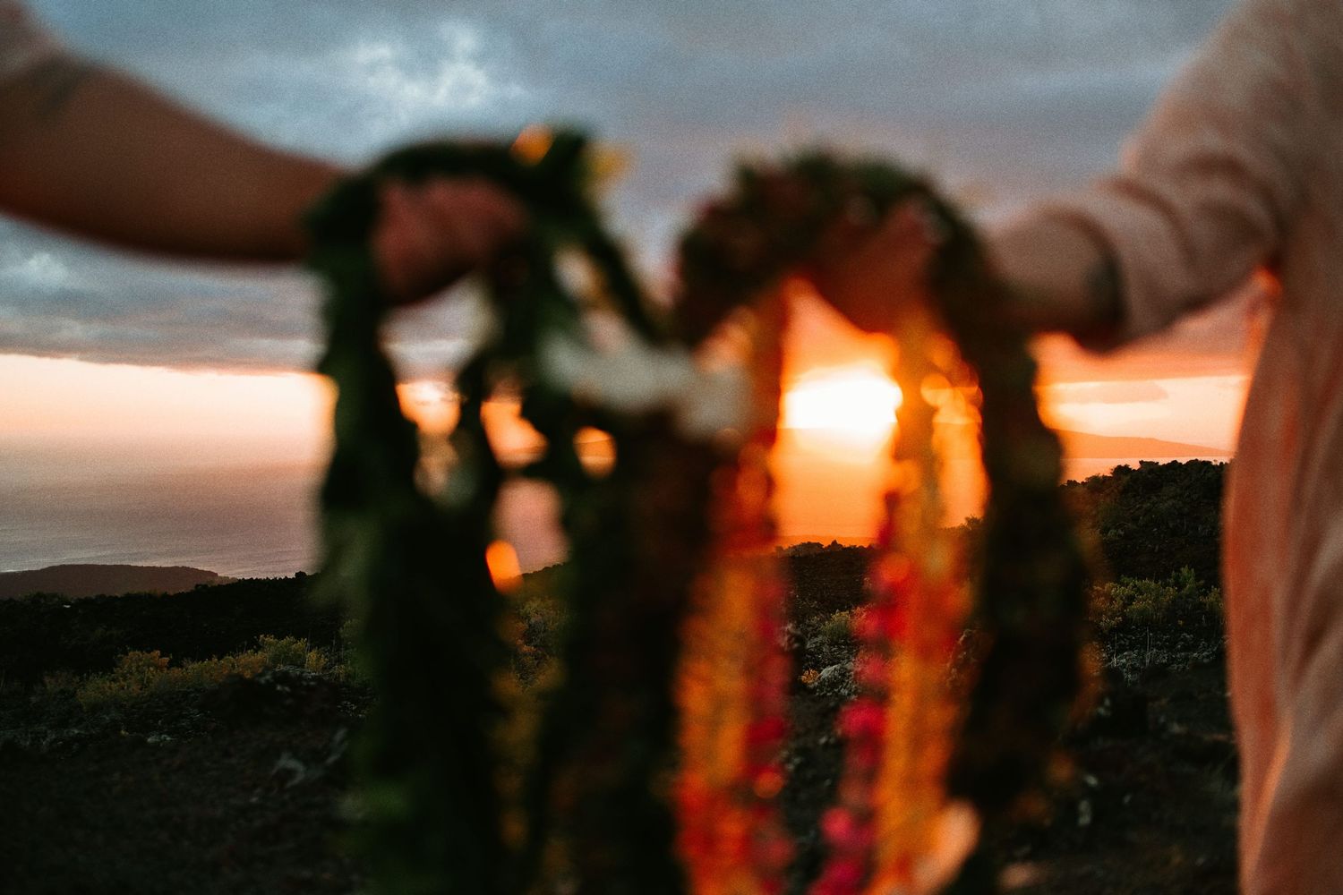 Couple holding floral leis at sunset with ocean views at a Hawaiian wedding ceremony.