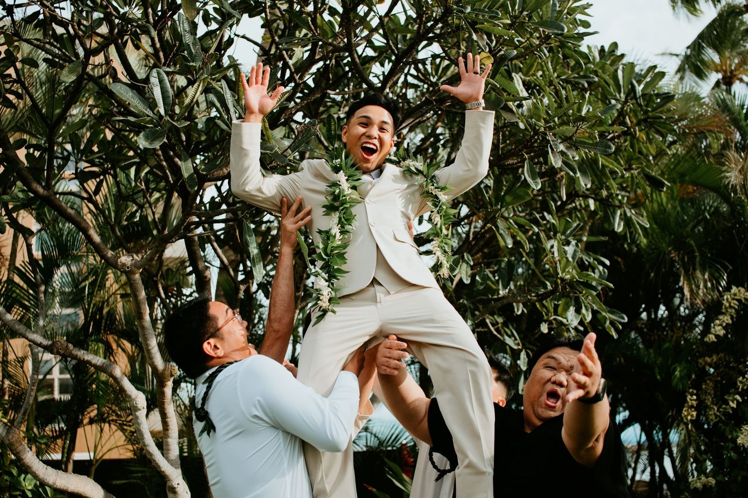 Groom in white suit lifted by groomsmen, celebrating joyfully outdoors with tropical trees in background.