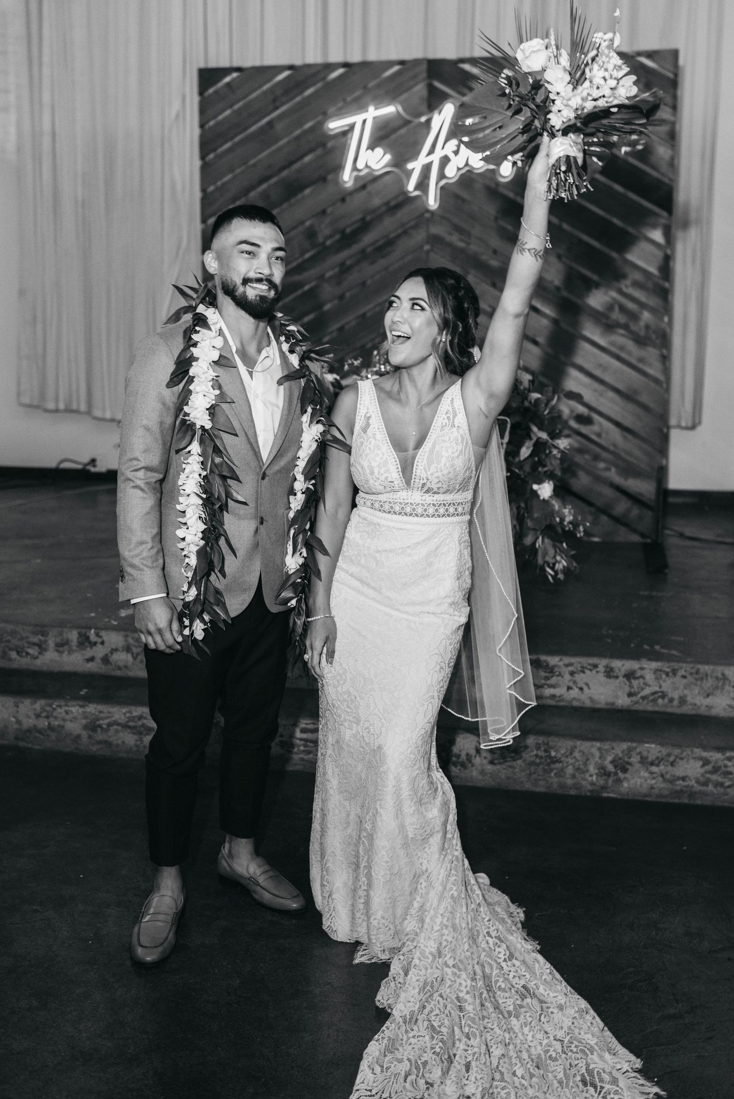 Black and white photo of a joyful newlywed couple celebrating, bride raising bouquet, groom wearing floral lei, neon sign backdrop.