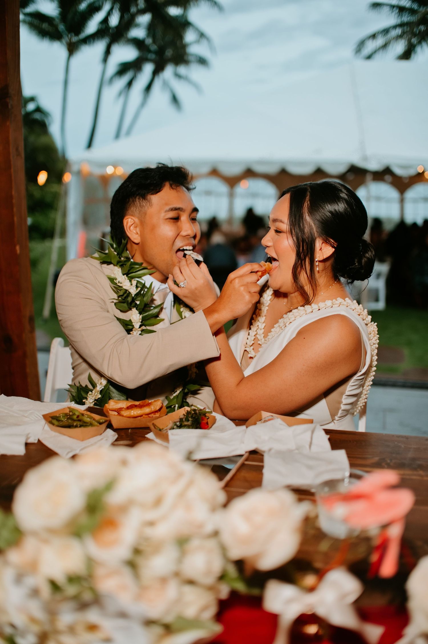 Newlywed couple feeding each other cake at outdoor Hawaiian wedding reception with floral leis and tropical decor.
