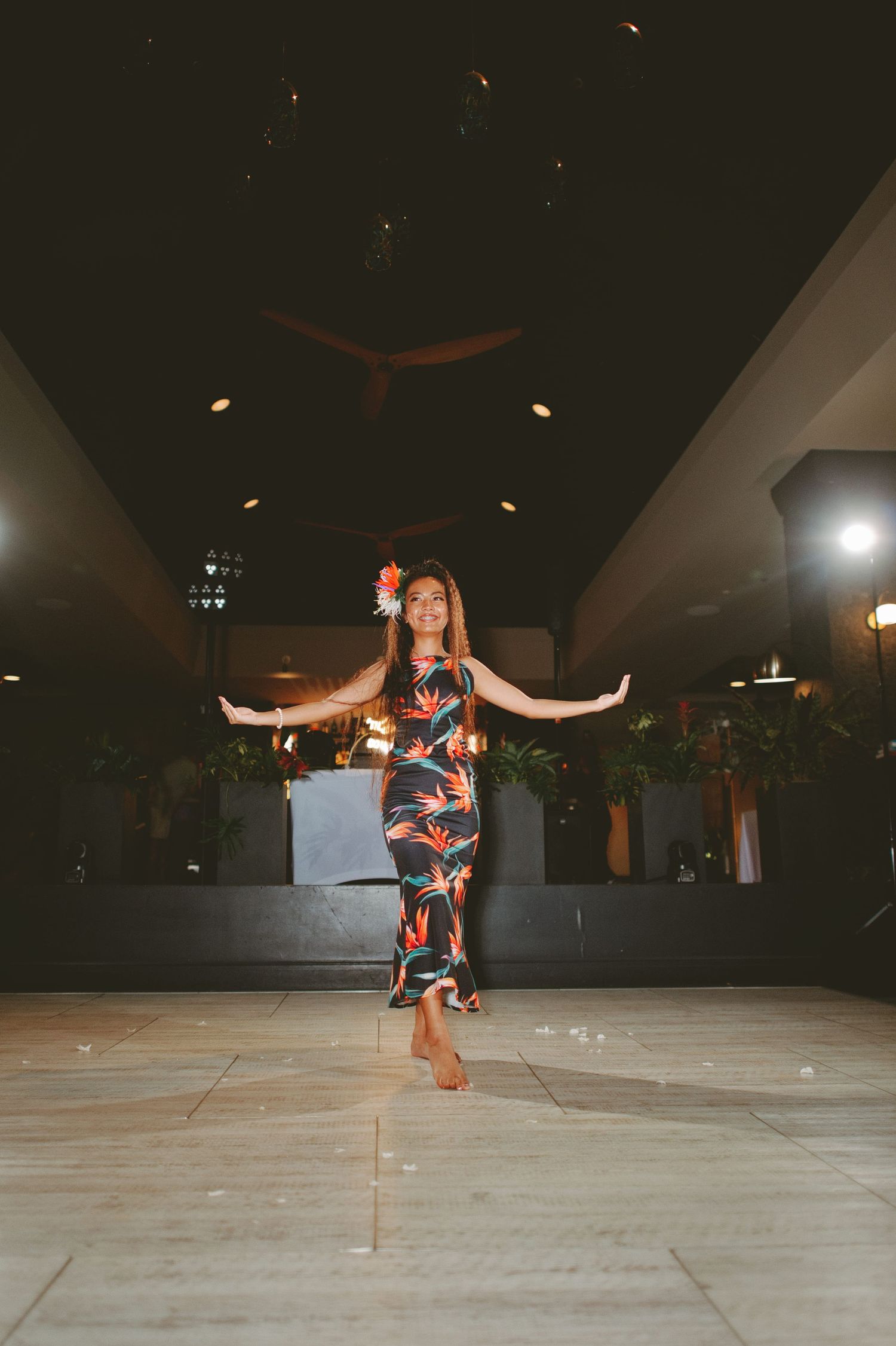 Woman in floral Hawaiian dress dancing barefoot in elegant indoor venue with tropical decor.