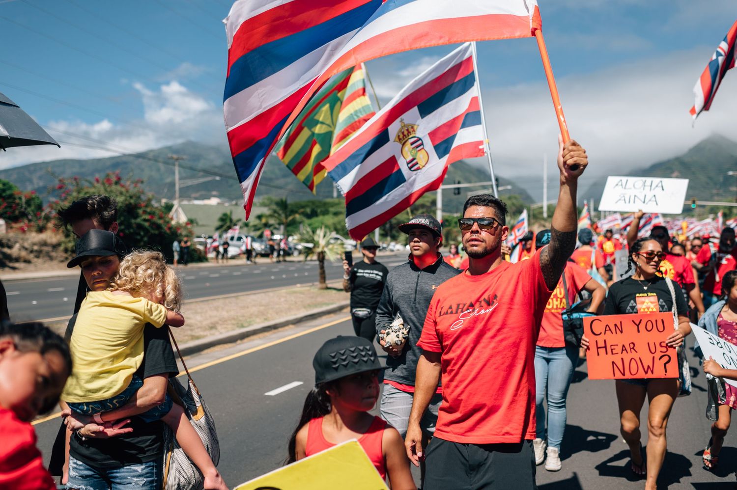 Demonstrators march with Hawaiian state flags and protest signs against mountain backdrop.