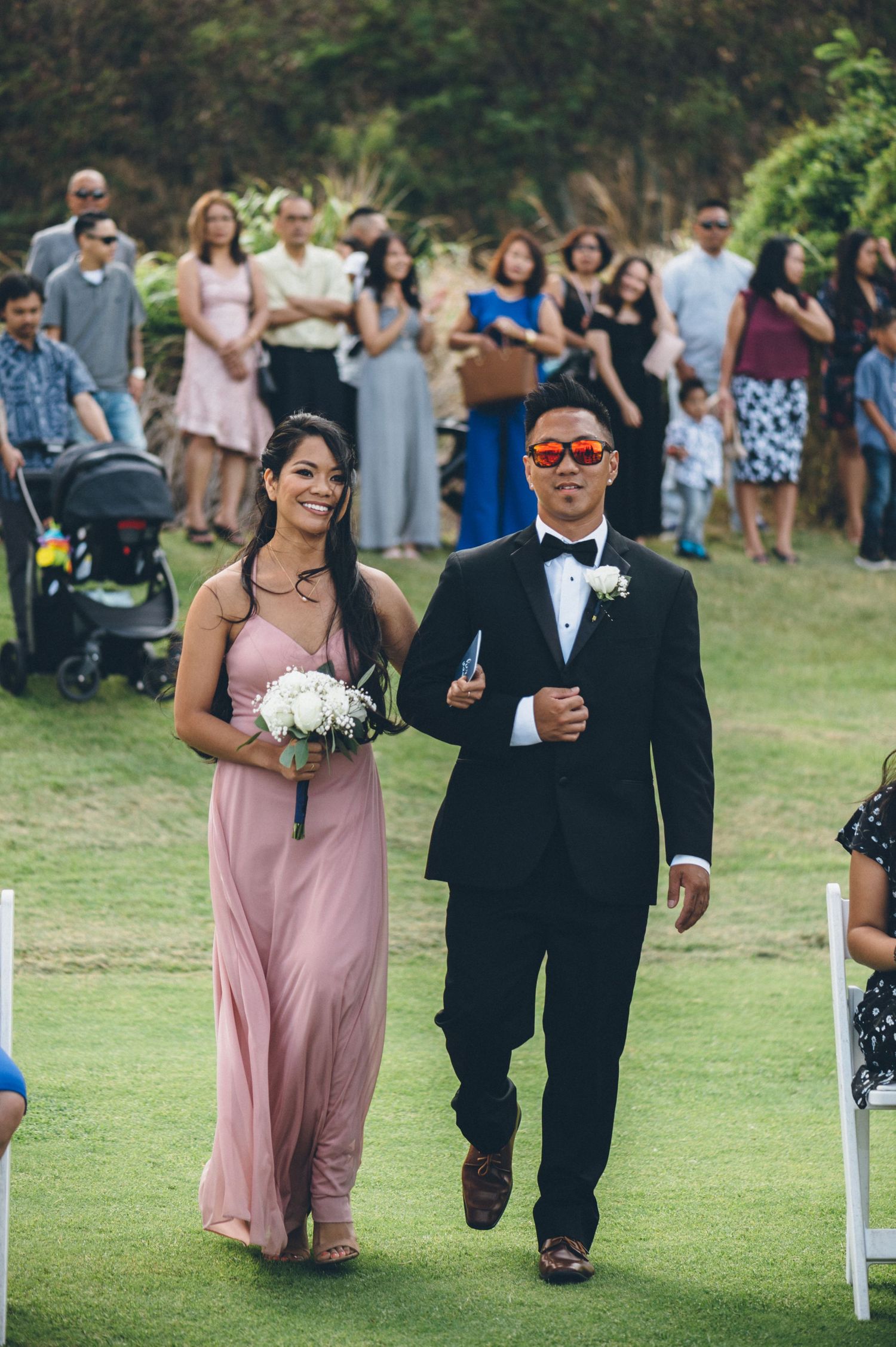Wedding party members walk down a grassy aisle with guests watching from both sides.