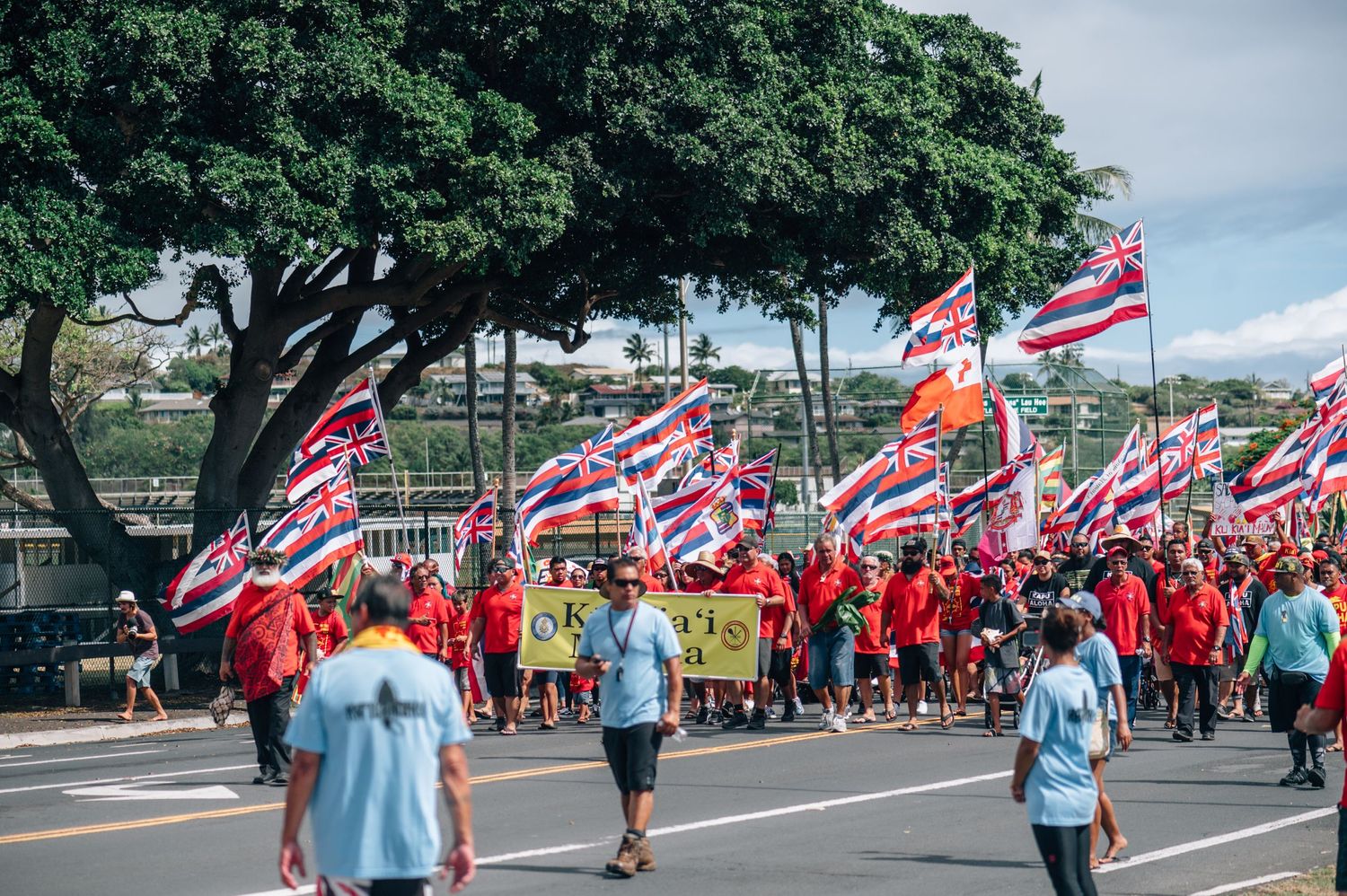 Protesters carrying Hawaiian flags march along a street while police vehicles follow behind.