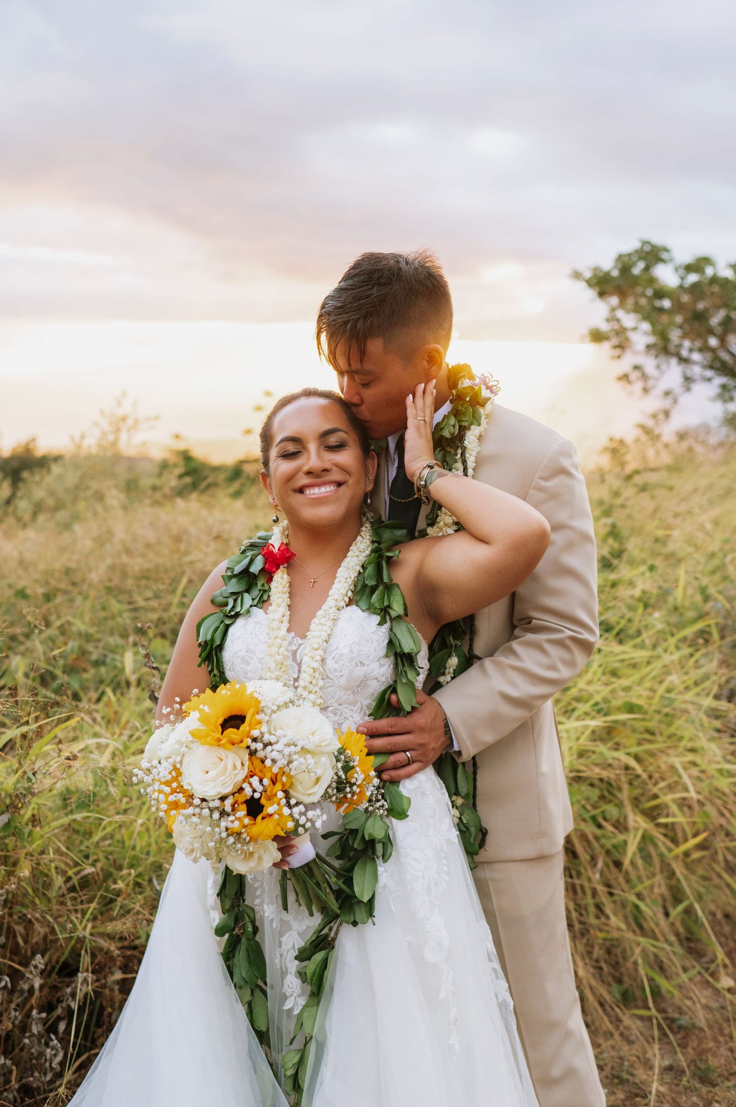 Bride and groom embrace at sunset in a field, wearing Hawaiian leis, as she holds a sunflower bouquet.