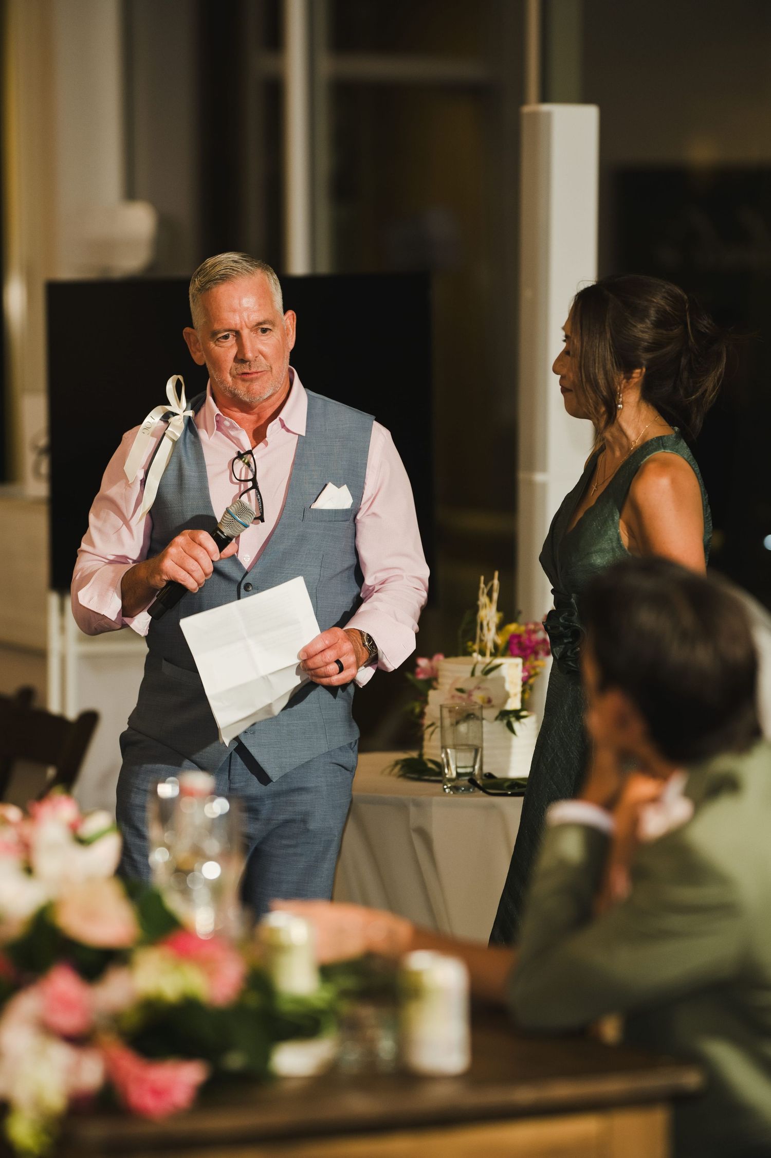 Man in gray vest holds microphone and paper, giving a speech at an elegant evening wedding reception.
