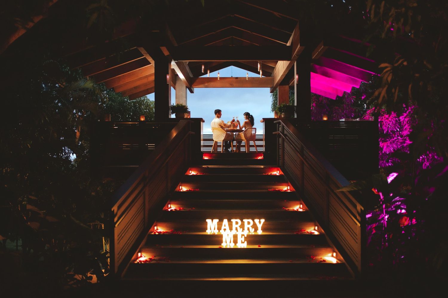 Romantic nighttime marriage proposal setup with glowing MARRY ME letters on staircase leading to couple under gazebo.