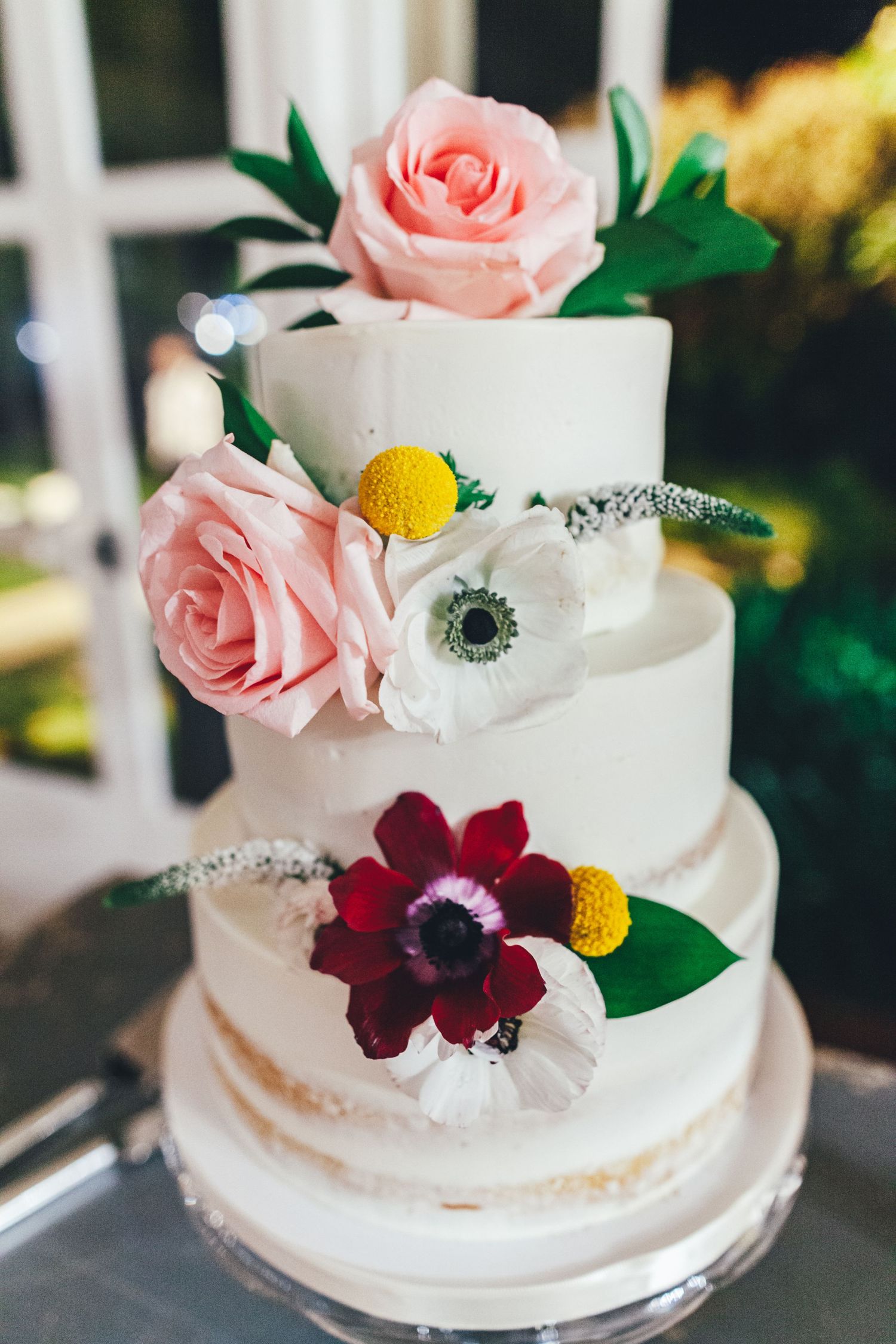 Elegant white wedding cake decorated with pink roses, burgundy flowers, and yellow craspedia blooms on display.