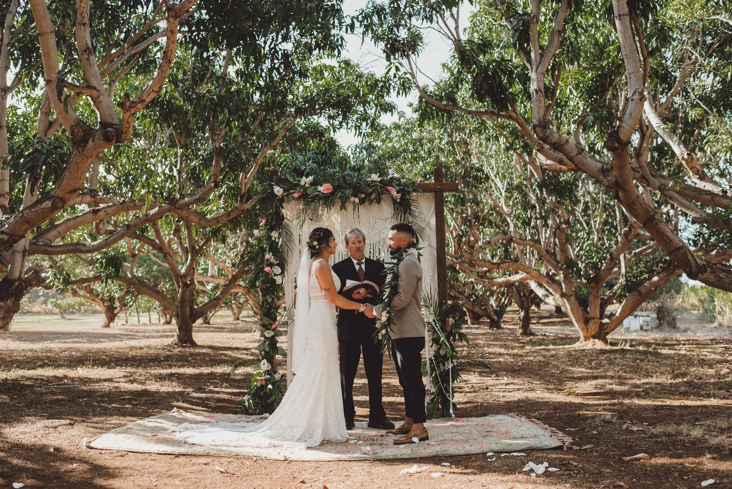 Outdoor wedding ceremony under eucalyptus trees with bride, groom, and officiant at a rustic floral arch.