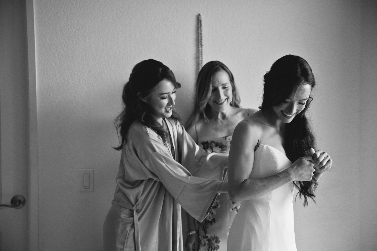 Black and white photo of bride being helped into wedding dress by two bridesmaids in robes.
