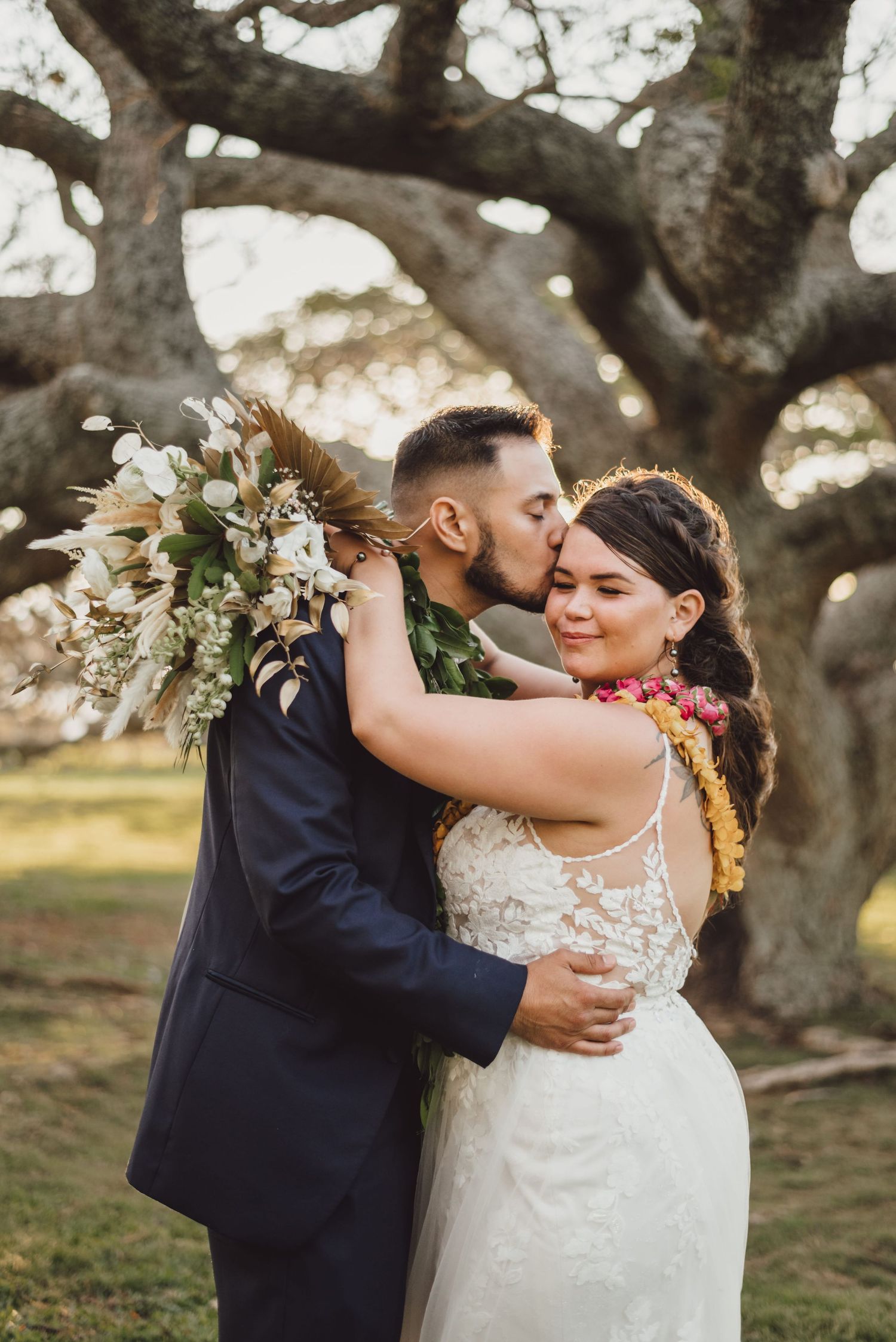 Bride and groom share a tender kiss under a large oak tree, bride holding a lush floral bouquet in a lace gown.