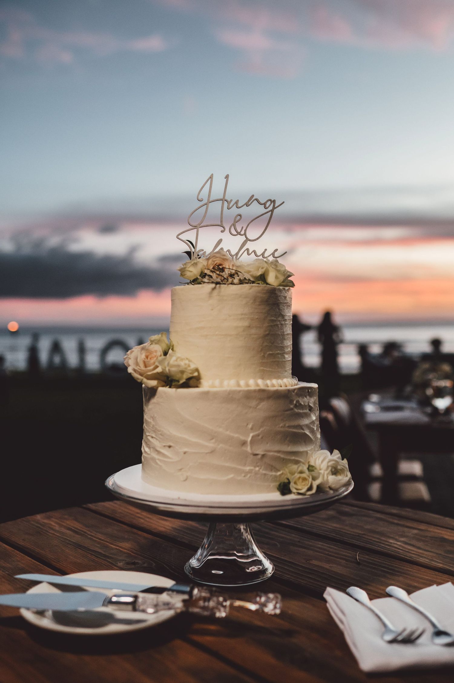 Two-tier white wedding cake with floral decor and custom topper, displayed outdoors at sunset near the ocean.