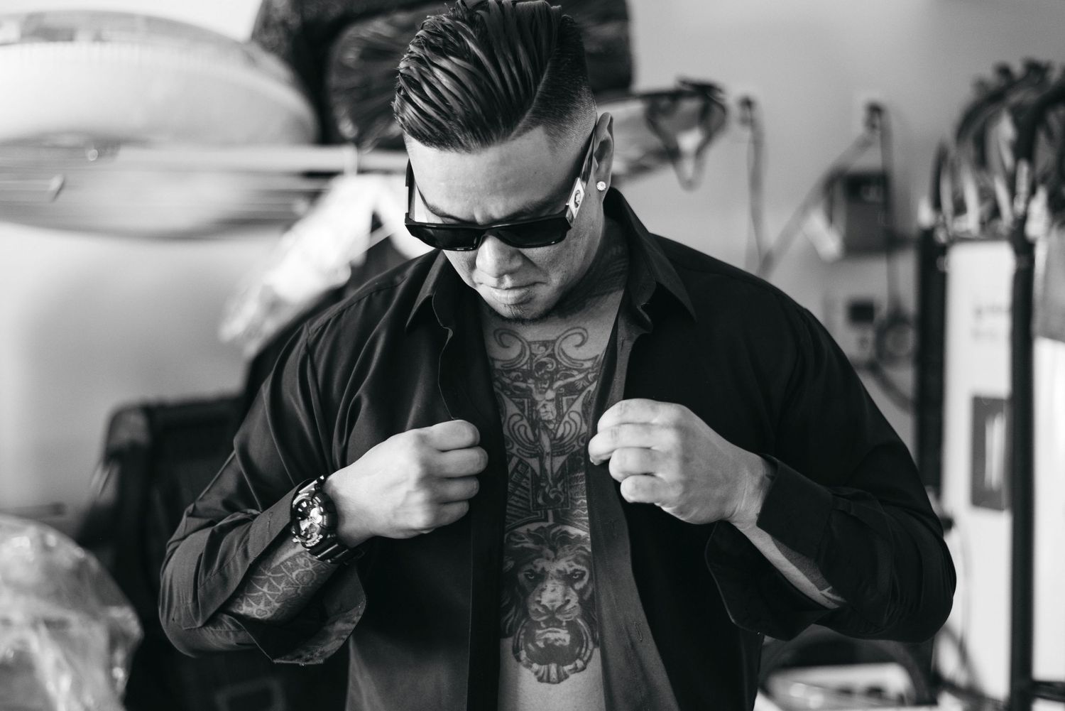 Tattooed man in black shirt and sunglasses posing with fists raised, showing chest tattoos in black and white photo.