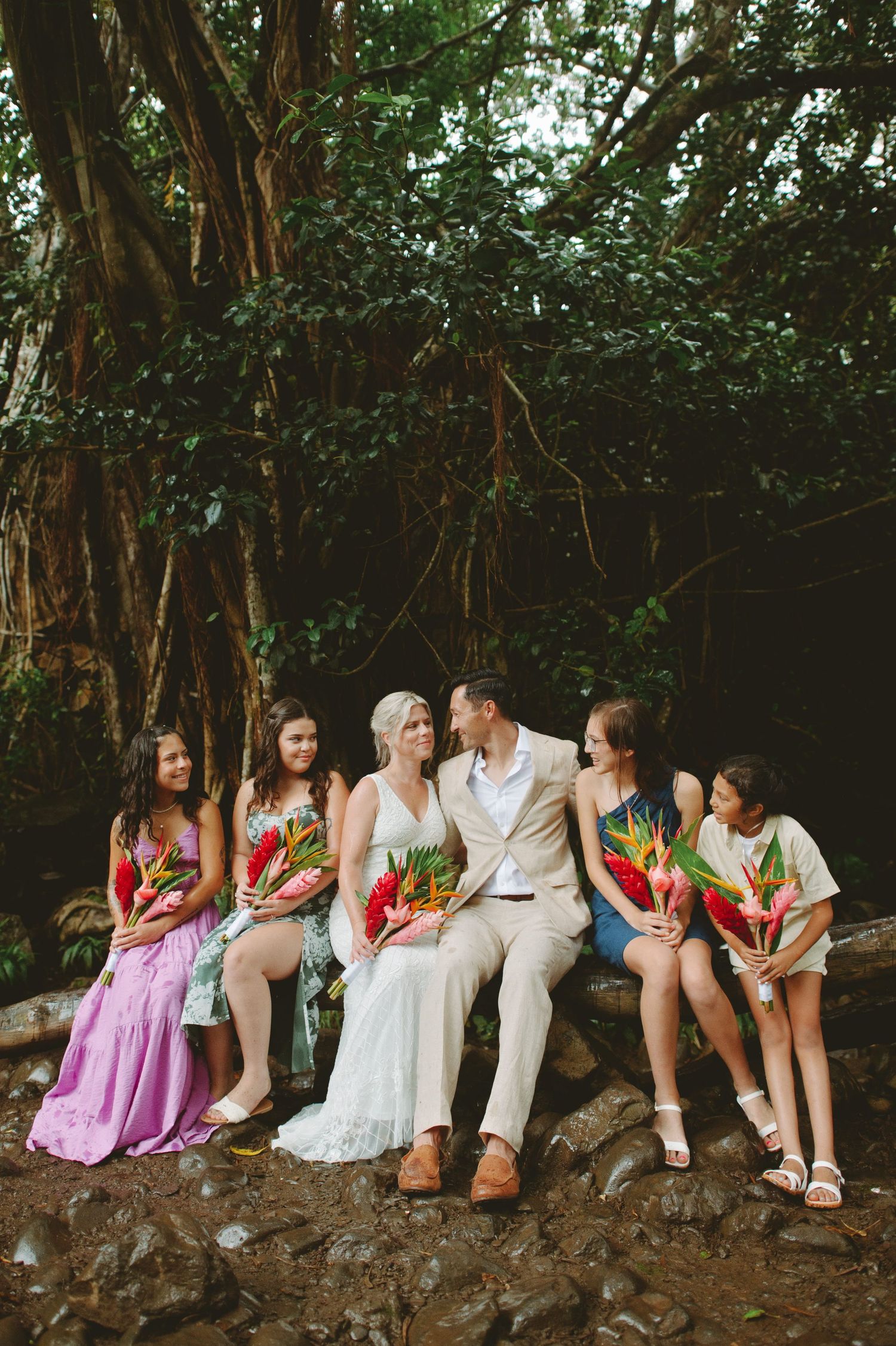 Bride and groom share a kiss surrounded by bridesmaids holding tropical flowers, seated on rocks in a lush garden setting.