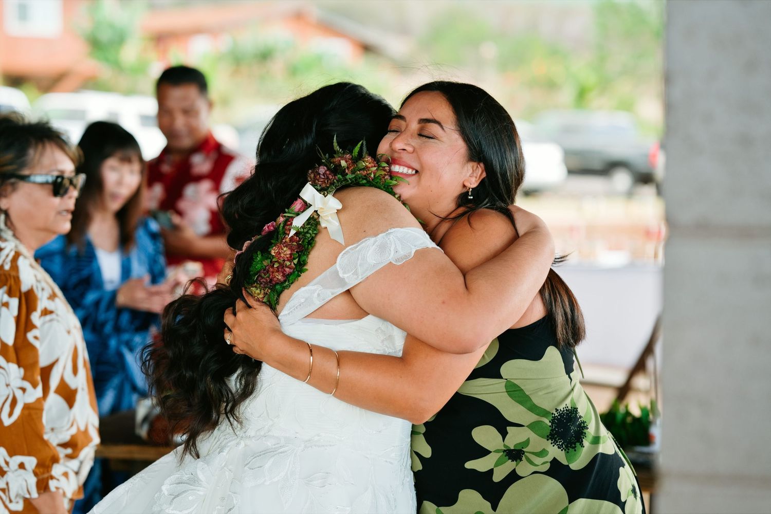 Bride in white lace dress shares a warm hug with a guest in a floral dress at an outdoor wedding reception.