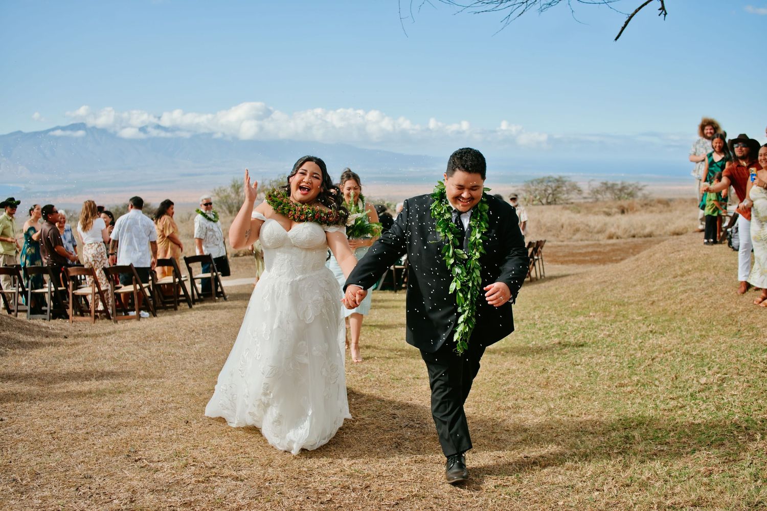 Joyful newlyweds wearing floral leis walk down outdoor aisle at Hawaiian wedding with mountain backdrop.