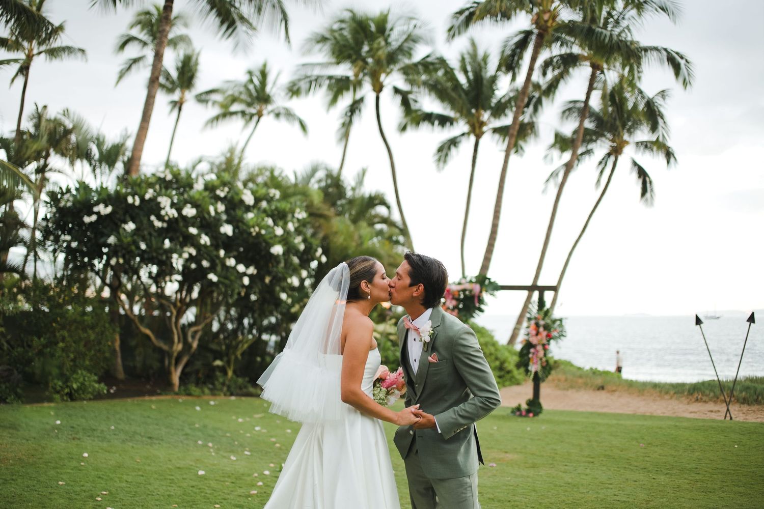 Bride and groom share a romantic kiss at an outdoor beach wedding ceremony surrounded by tropical palm trees in Maui.