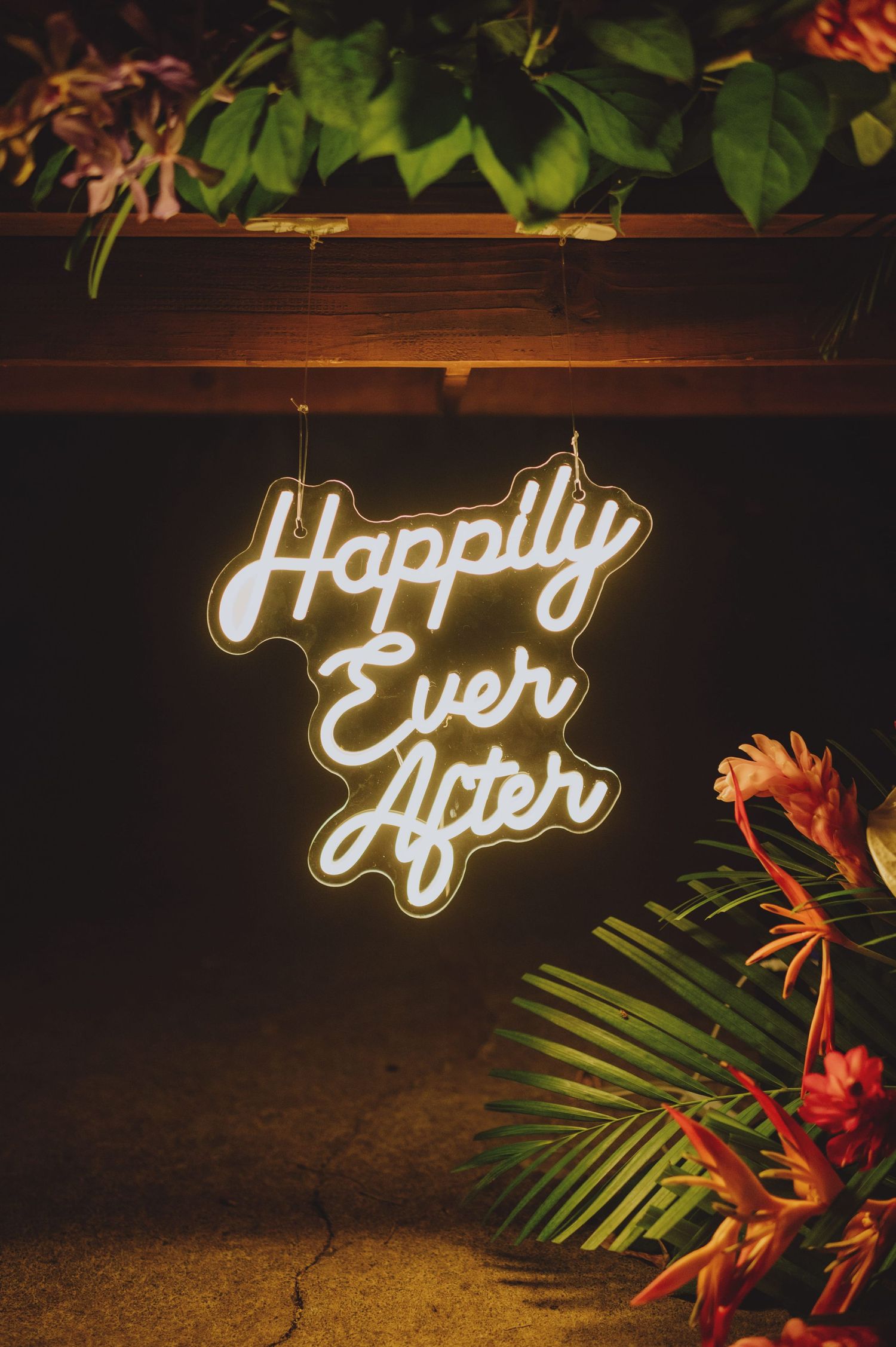 Glowing neon sign reading Happily Ever After hanging under wooden shelf with tropical flowers and greenery.