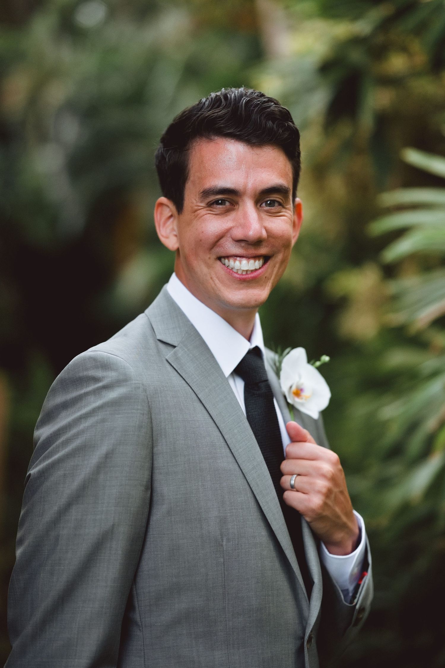 Smiling groom in gray suit with black tie and white orchid boutonniere posing outdoors among lush green tropical foliage.