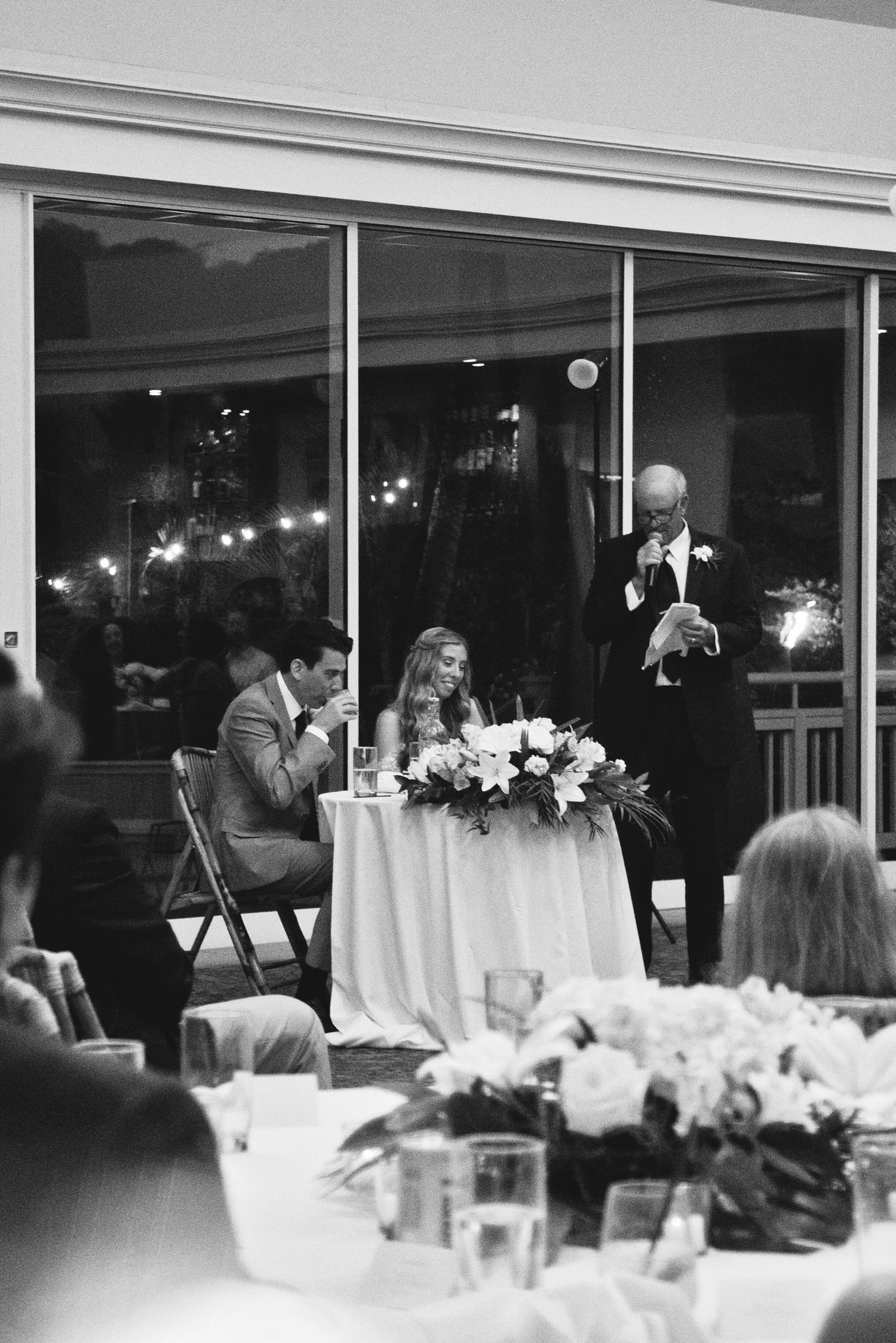 Black and white wedding reception photo showing bride and groom at sweetheart table during a heartfelt toast speech.