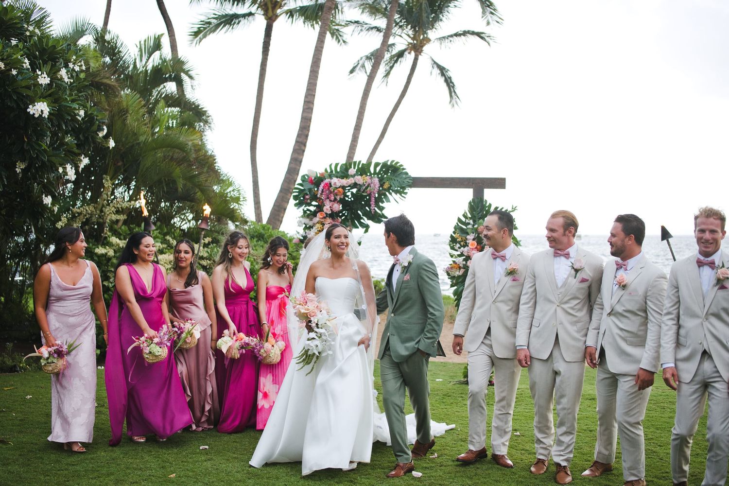 Tropical outdoor wedding party with bridesmaids in pink dresses and groomsmen in tan suits under palm trees in Hawaii.