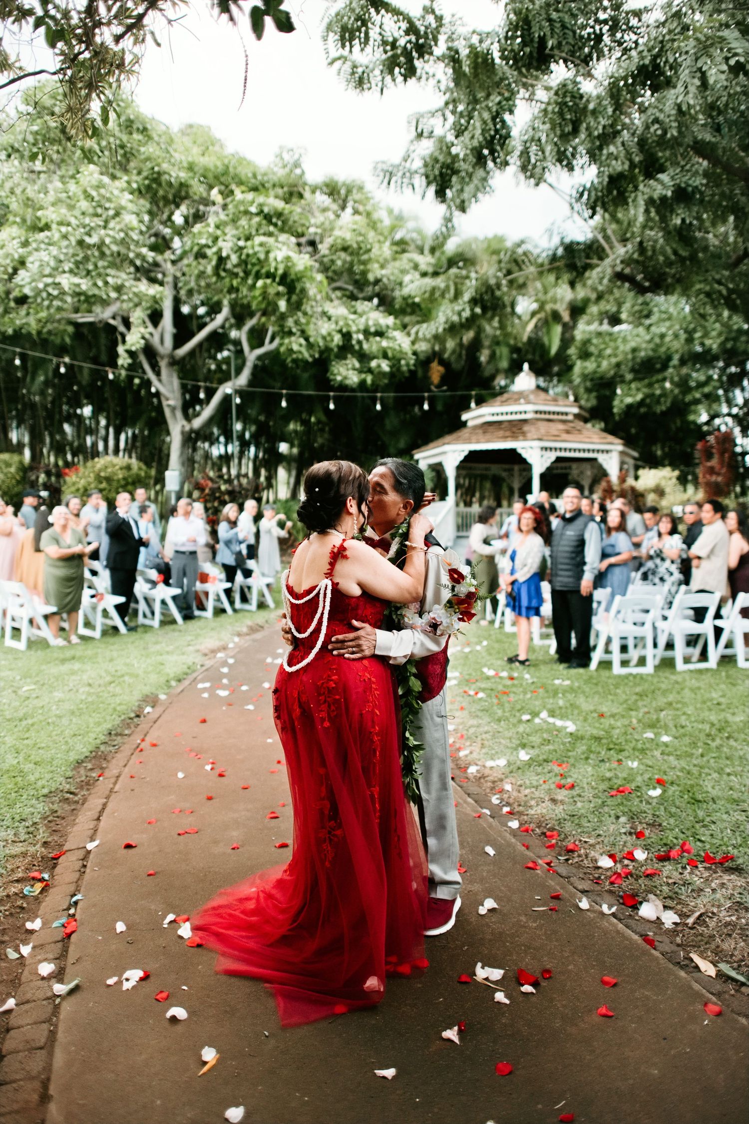 Couple in red wedding attire share first kiss on petal-strewn aisle with gazebo and guests in background.