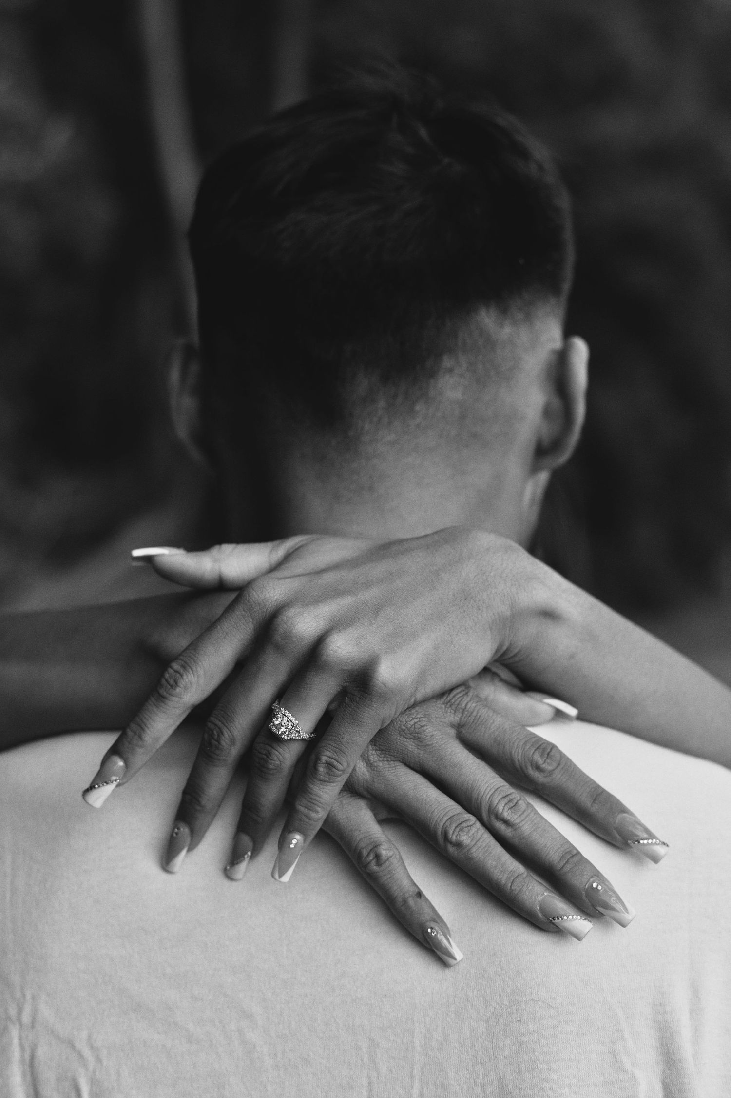 Close-up black and white photo of womans hands with engagement ring resting on mans back.