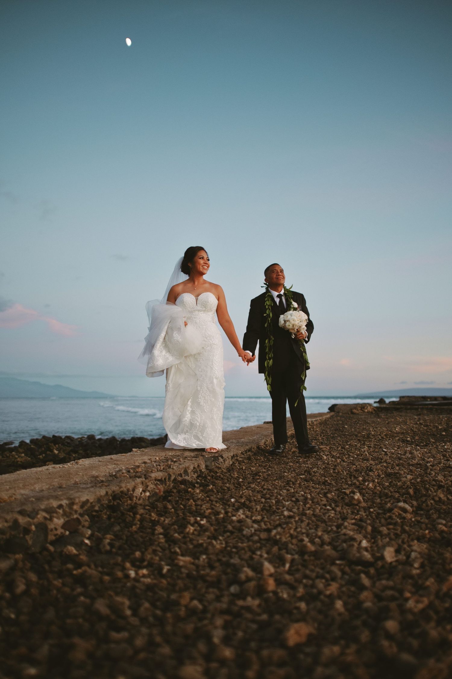 Bride and groom holding hands on rocky shoreline at dusk with ocean and moon backdrop.