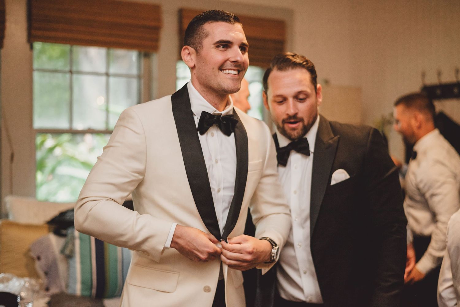 Groom in ivory tuxedo jacket with black lapels smiling as groomsman helps him button up before wedding ceremony.