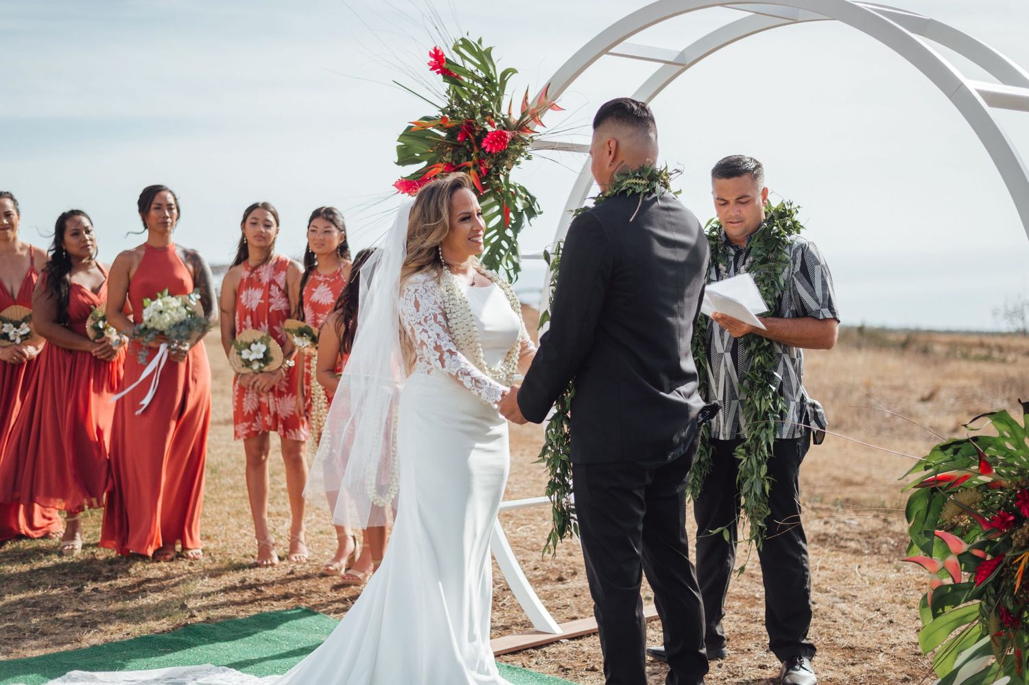 Outdoor Hawaiian wedding ceremony with bride in white lace gown, groom, officiant, and bridesmaids in red dresses under floral arch.