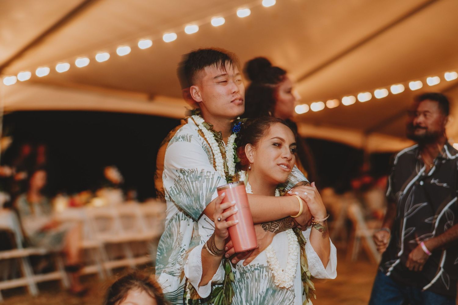 Couple in floral leis embracing at a festive outdoor wedding reception with string lights.