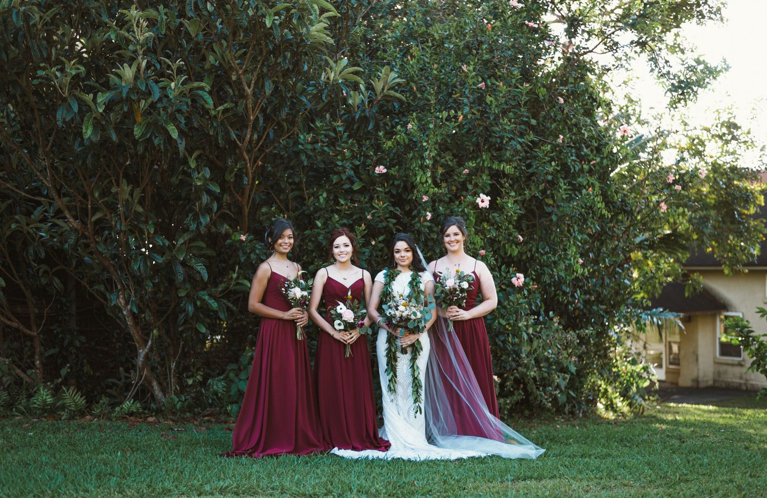 Bride in white gown with three bridesmaids in burgundy dresses holding bouquets outdoors near lush green trees.