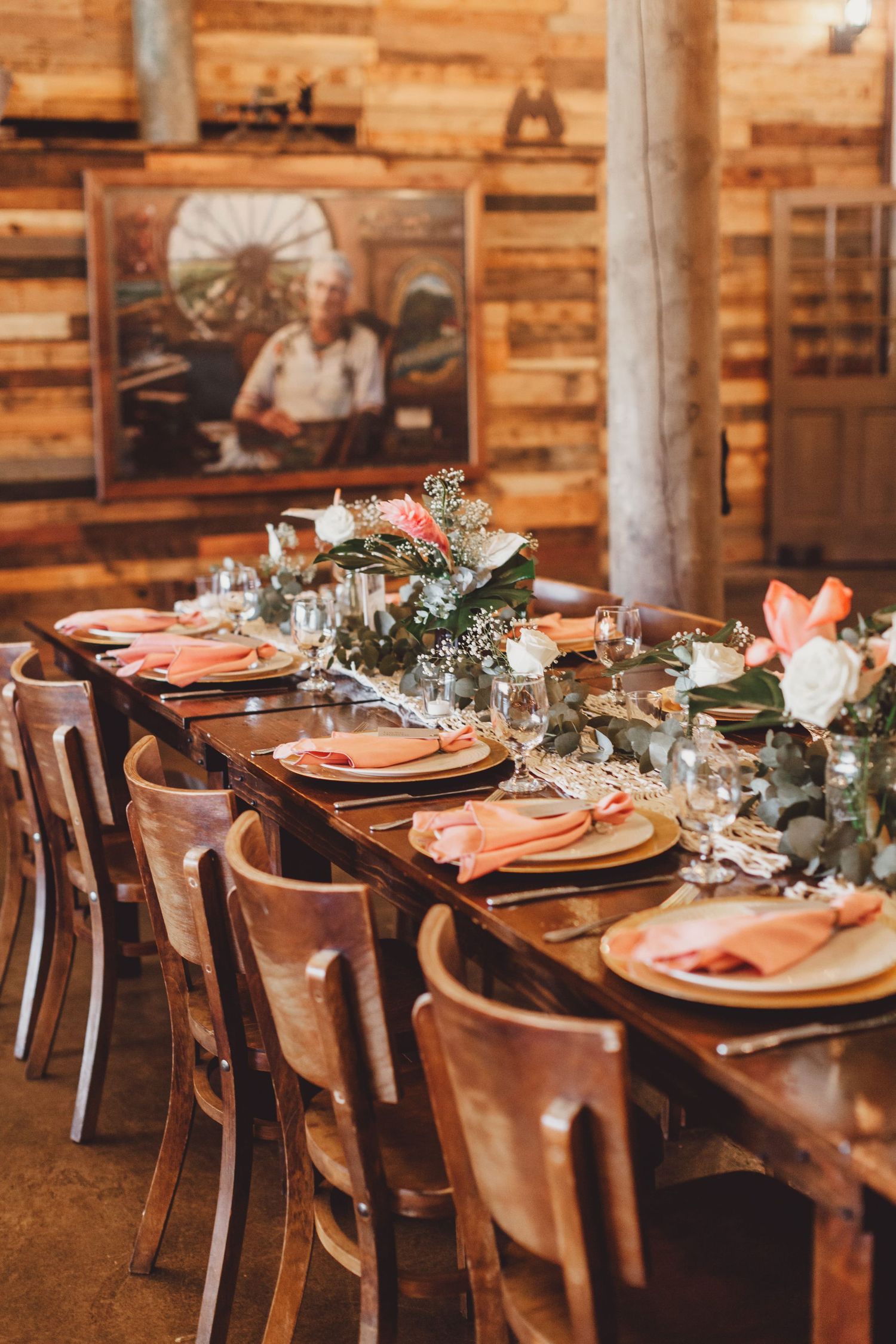 Rustic barn wedding reception with long wooden table, coral napkins, floral centerpieces, and vintage portrait on wood-paneled wall.