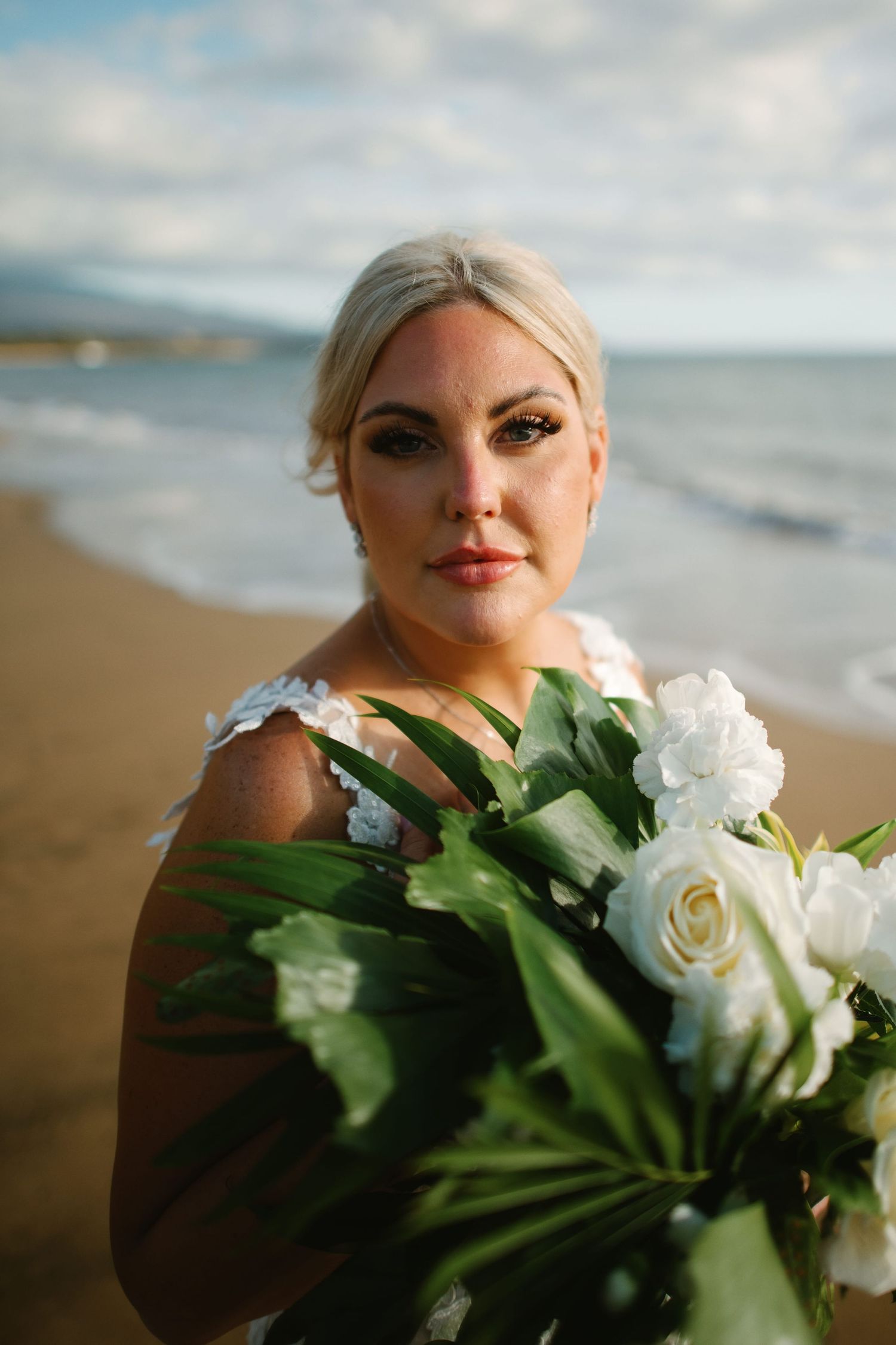 Bride holding white roses and tropical greenery bouquet on sandy beach, wearing white gown with ocean backdrop.