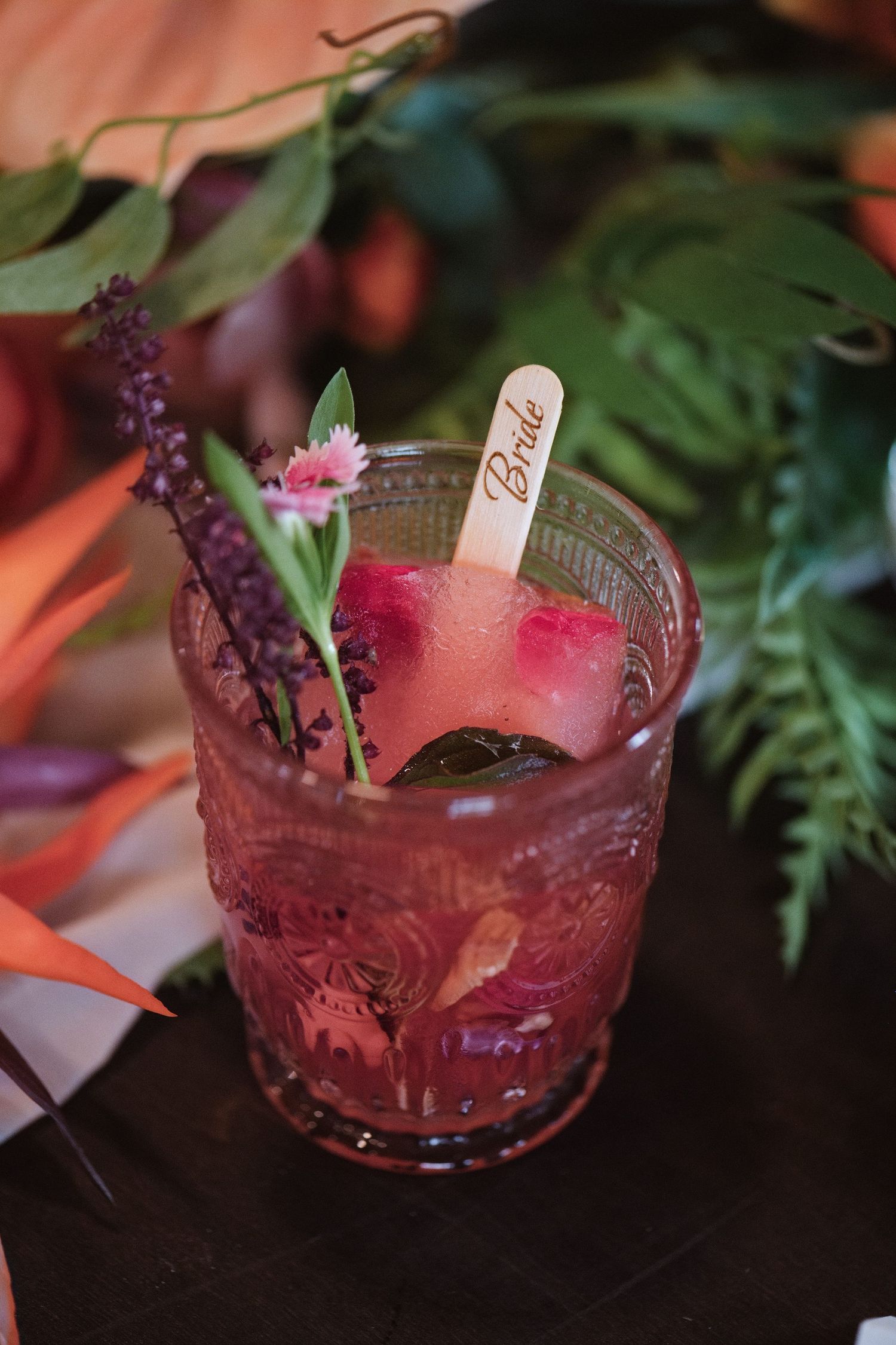 A bride's cocktail garnished with fresh flowers, lavender, and a wooden bride pick in a vintage glass.