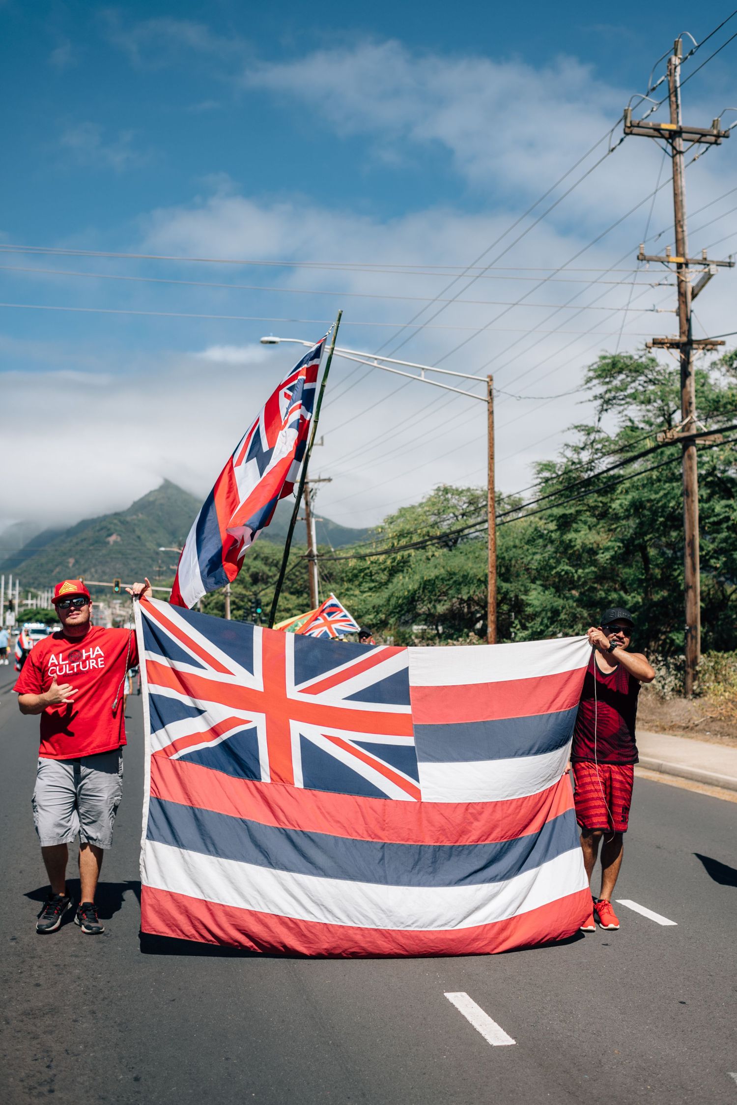 Group displays large Hawaiian state flag across street with mountains and power lines in background.