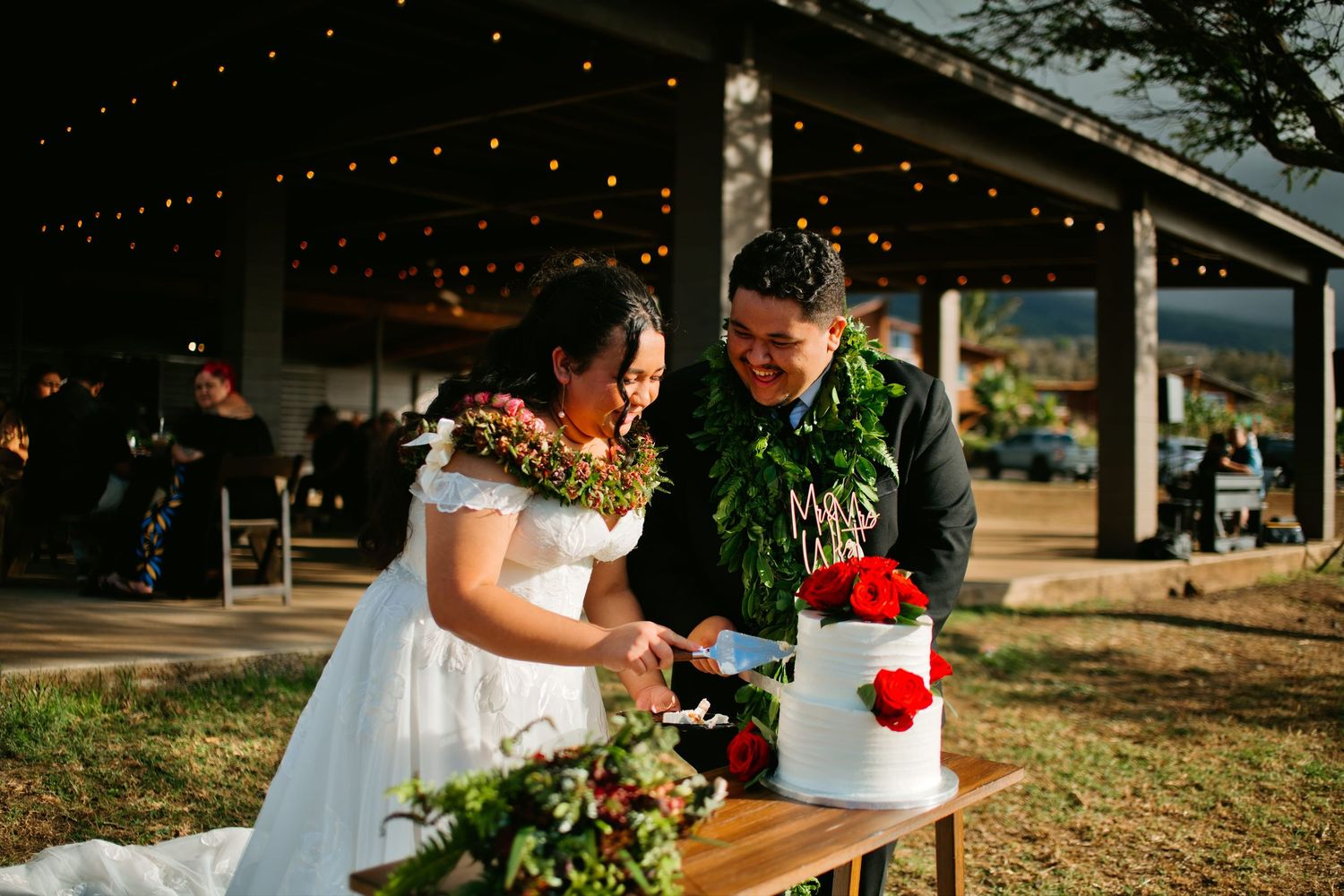Hawaiian wedding couple cutting white cake outdoors under string lights, wearing floral leis at rustic venue reception.
