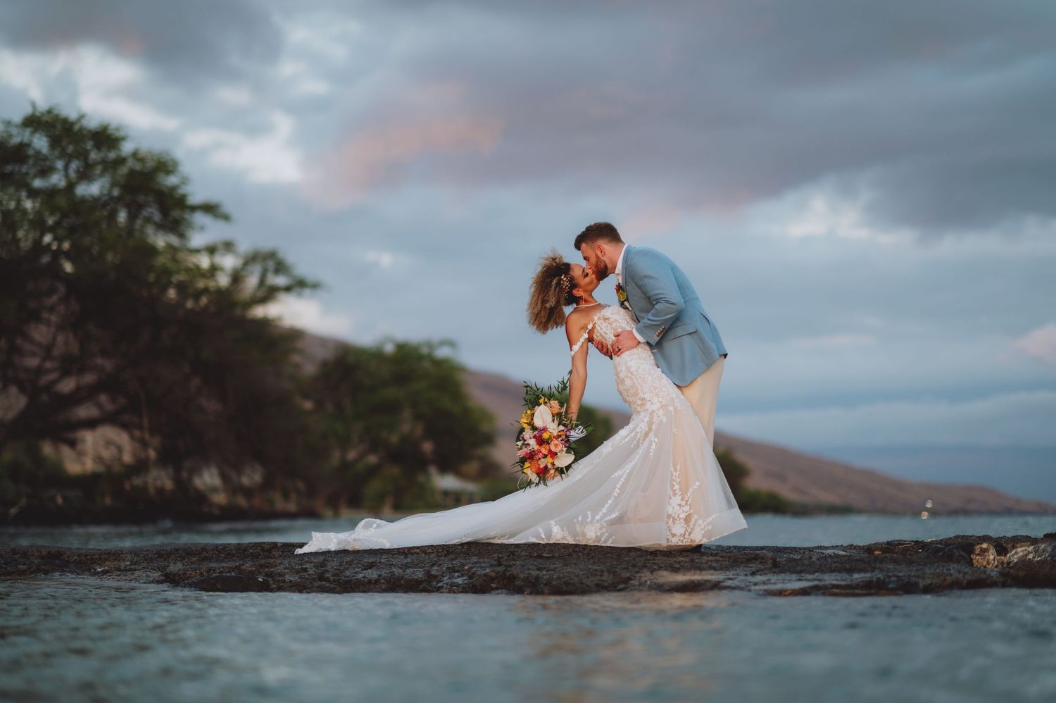 Bride and groom share a romantic dip kiss on a rocky shoreline at sunset in Hawaii, with mountains in the background.