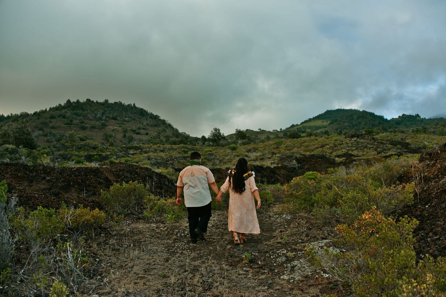 Couple holds hands walking through a scenic volcanic landscape with green hills and cloudy skies.