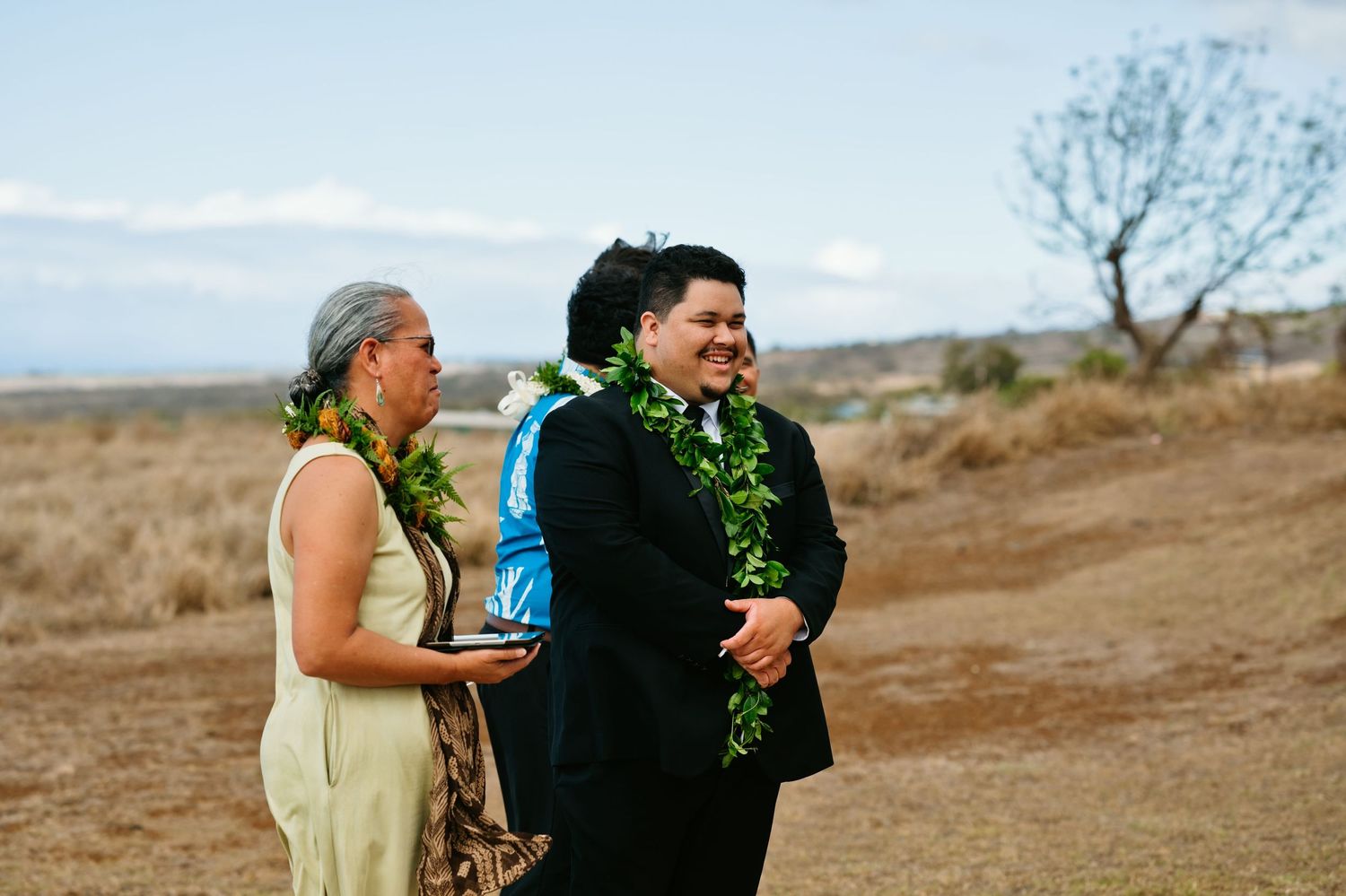 Two people in formal attire with lei garlands laugh during an outdoor Hawaiian wedding ceremony on a dry field.