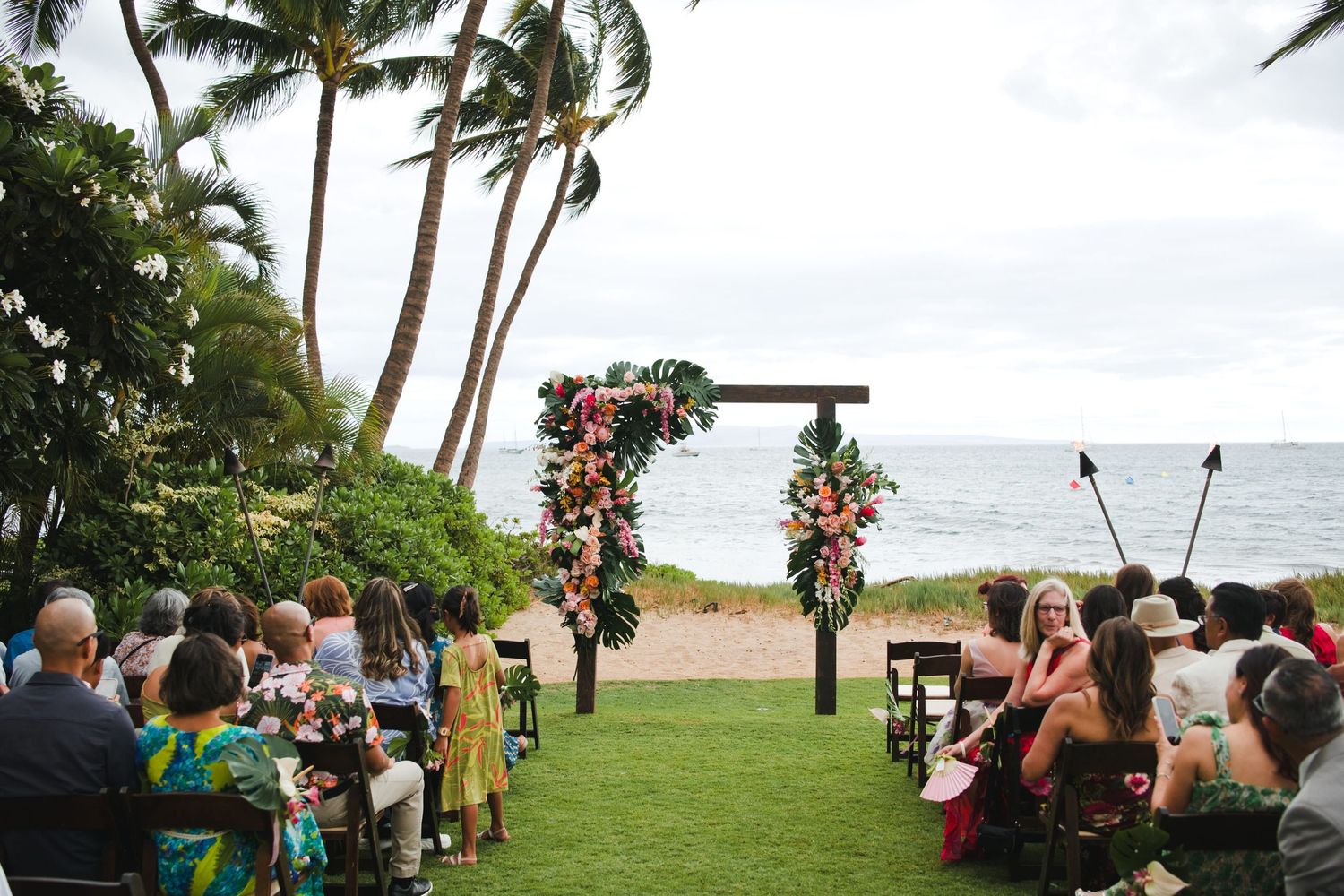 Outdoor beach wedding ceremony with floral arch, palm trees, and ocean backdrop in Hawaii.